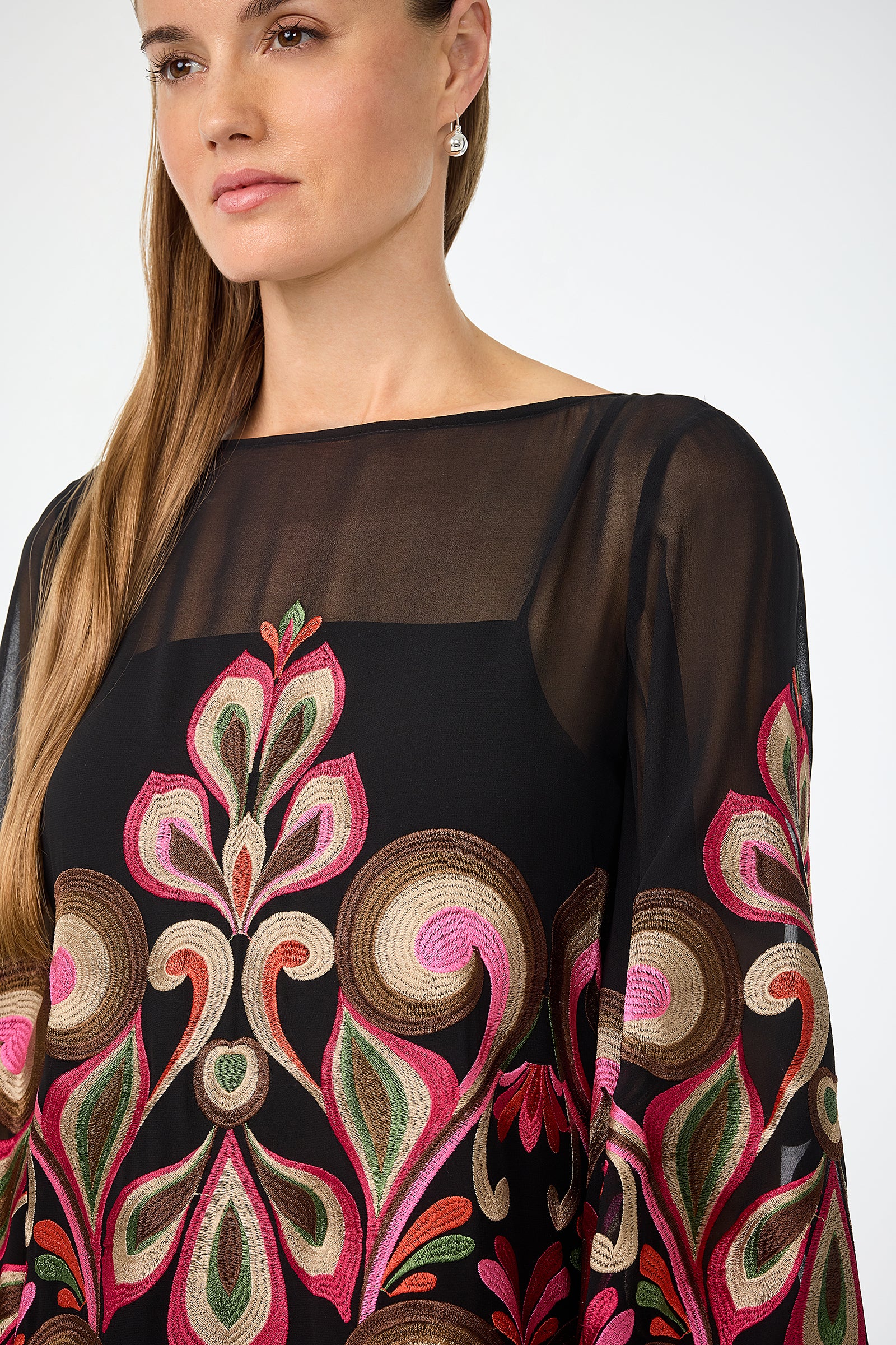 A woman with long, straight hair wears the Salim Embroidered Mini Dress—a sheer black piece with ornate pink, gold, and green embroidery. She has a neutral expression and small drop earrings against a plain white background.