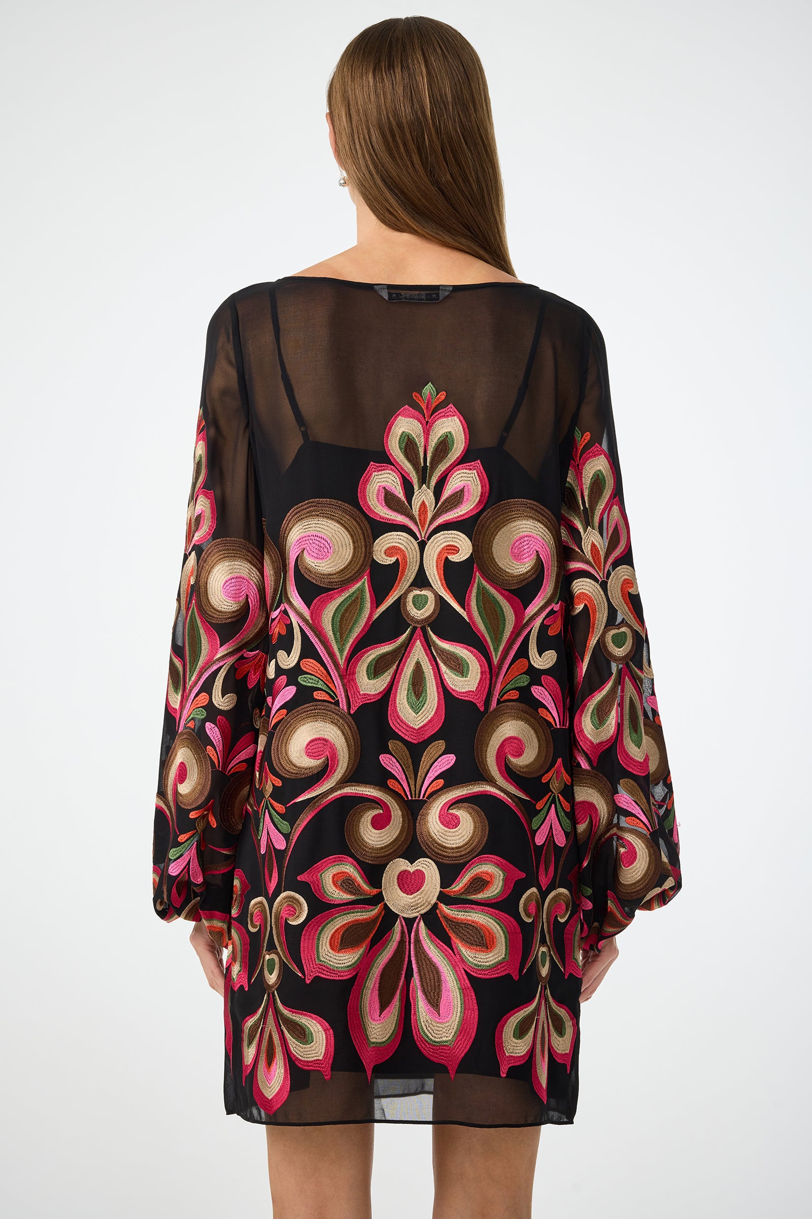 A woman with straight brown hair, facing away from the camera against a plain background, wears the Salim Embroidered Mini Dress featuring sheer black fabric and vibrant pink, red, green, and gold floral patterns on the back and sleeves.