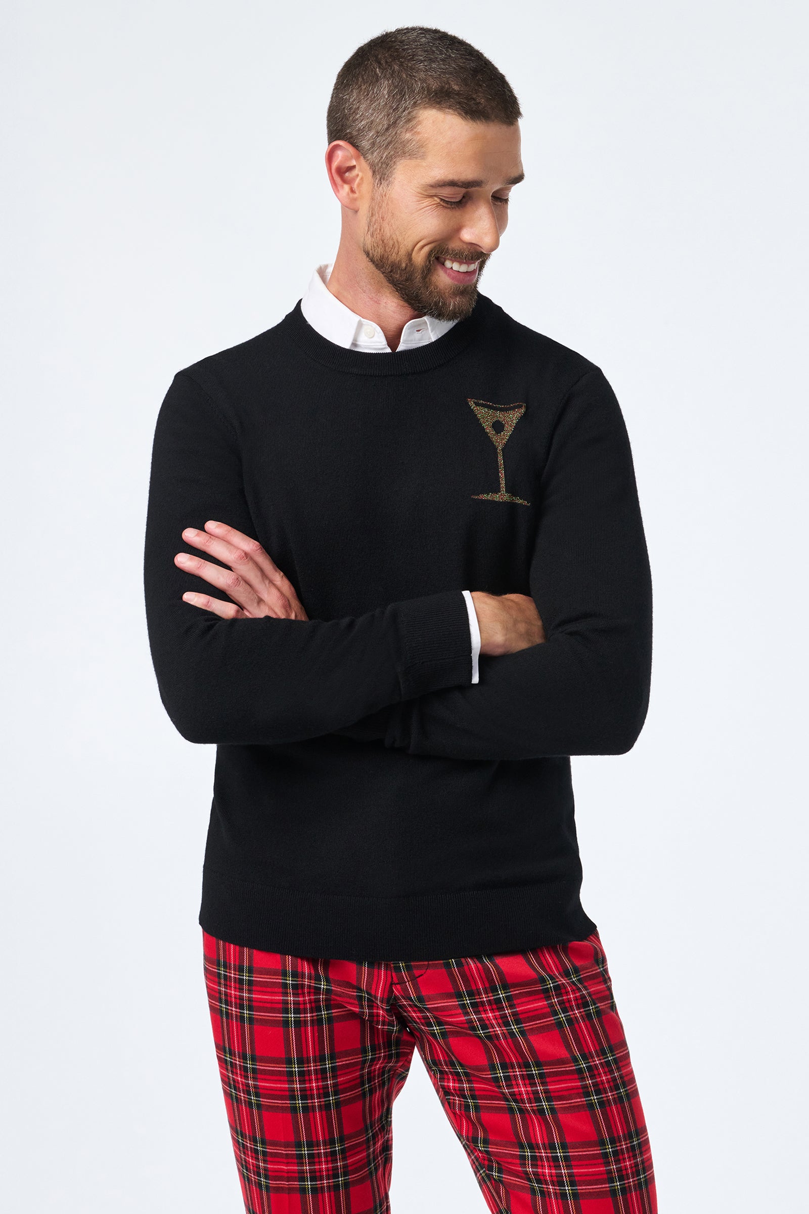 A man with short hair and a trimmed beard smiles with arms crossed, wearing the Martini Crewneck—a black sweater featuring a martini glass graphic—and red plaid pants.