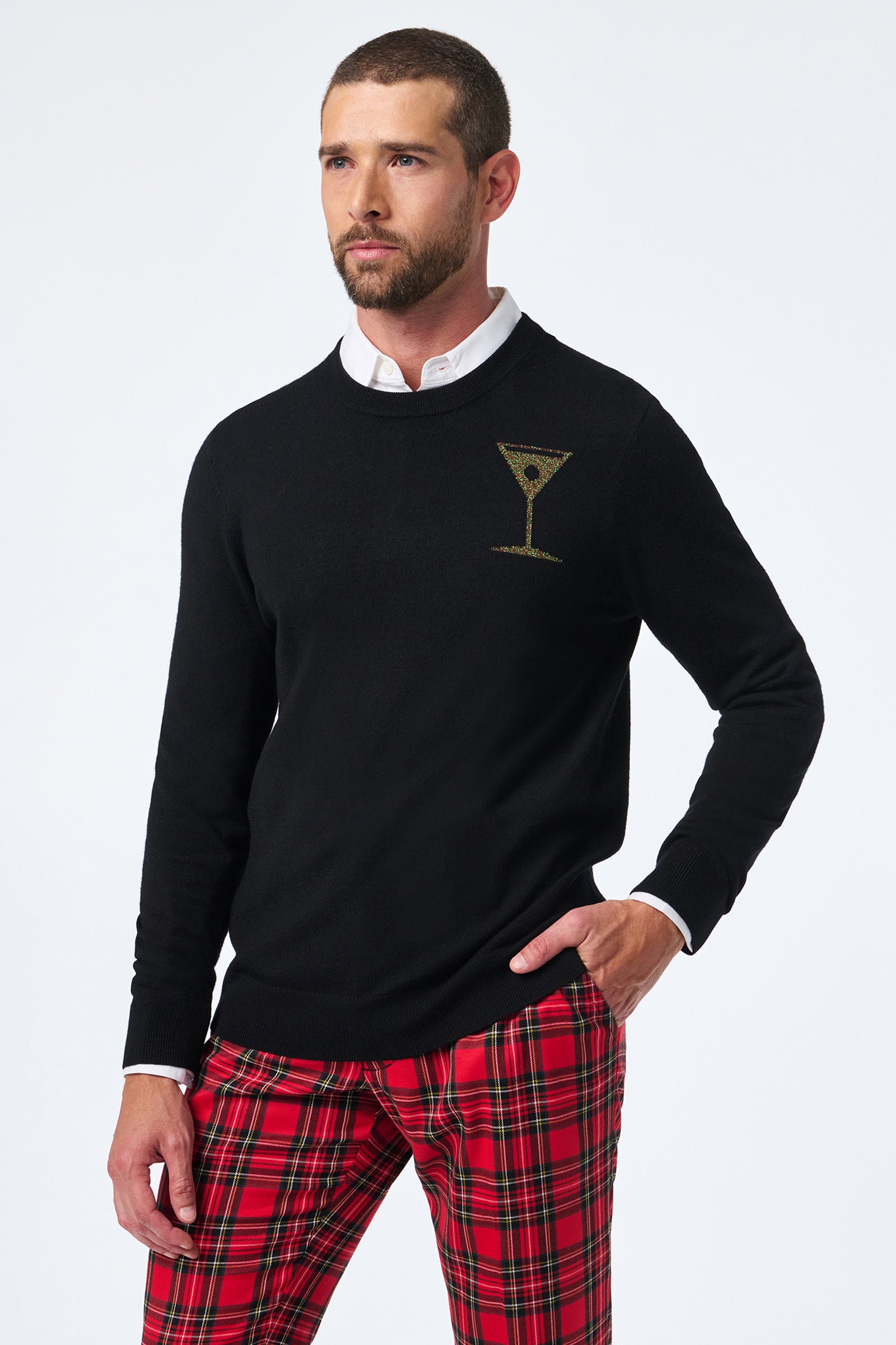 A man with short hair and a beard wears the Martini Crewneck—black with a martini glass design—over a white collared shirt and red plaid pants, standing against a plain light background.