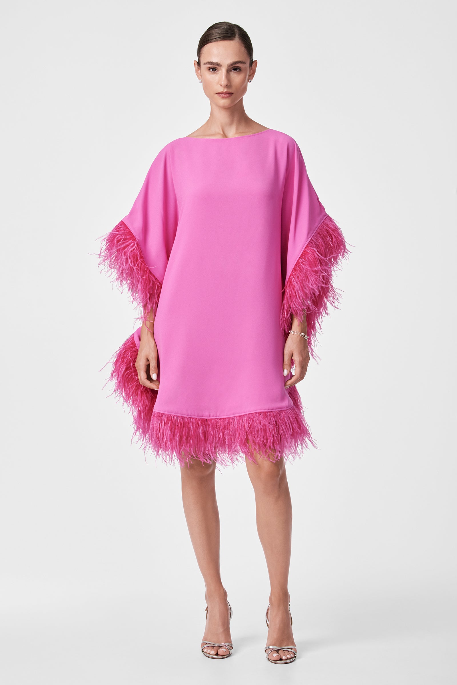 A woman wears the Mini Neena Caftan Dress, a loose-fitting, bright pink dress with feather trim on the sleeves and hem, paired with silver high-heeled sandals. Her hair is slicked back as she stands against a plain white background.