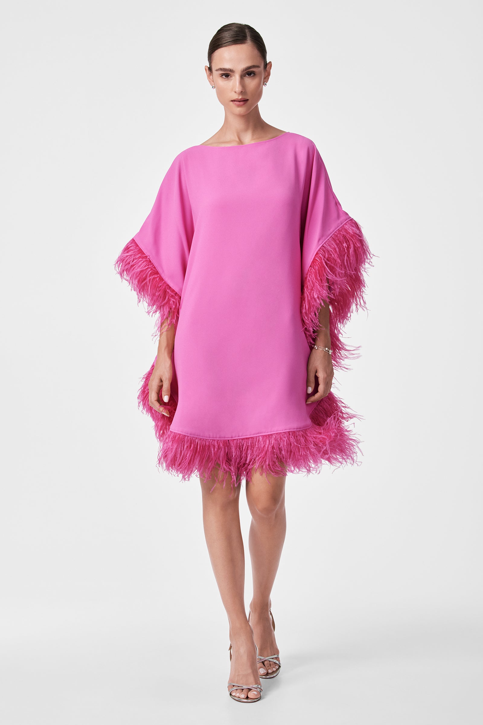 A woman stands against a plain white background wearing the Mini Neena Caftan Dress, a loose-fitting bright pink style with feathered trim on the sleeves and hem, paired with silver high-heeled sandals.