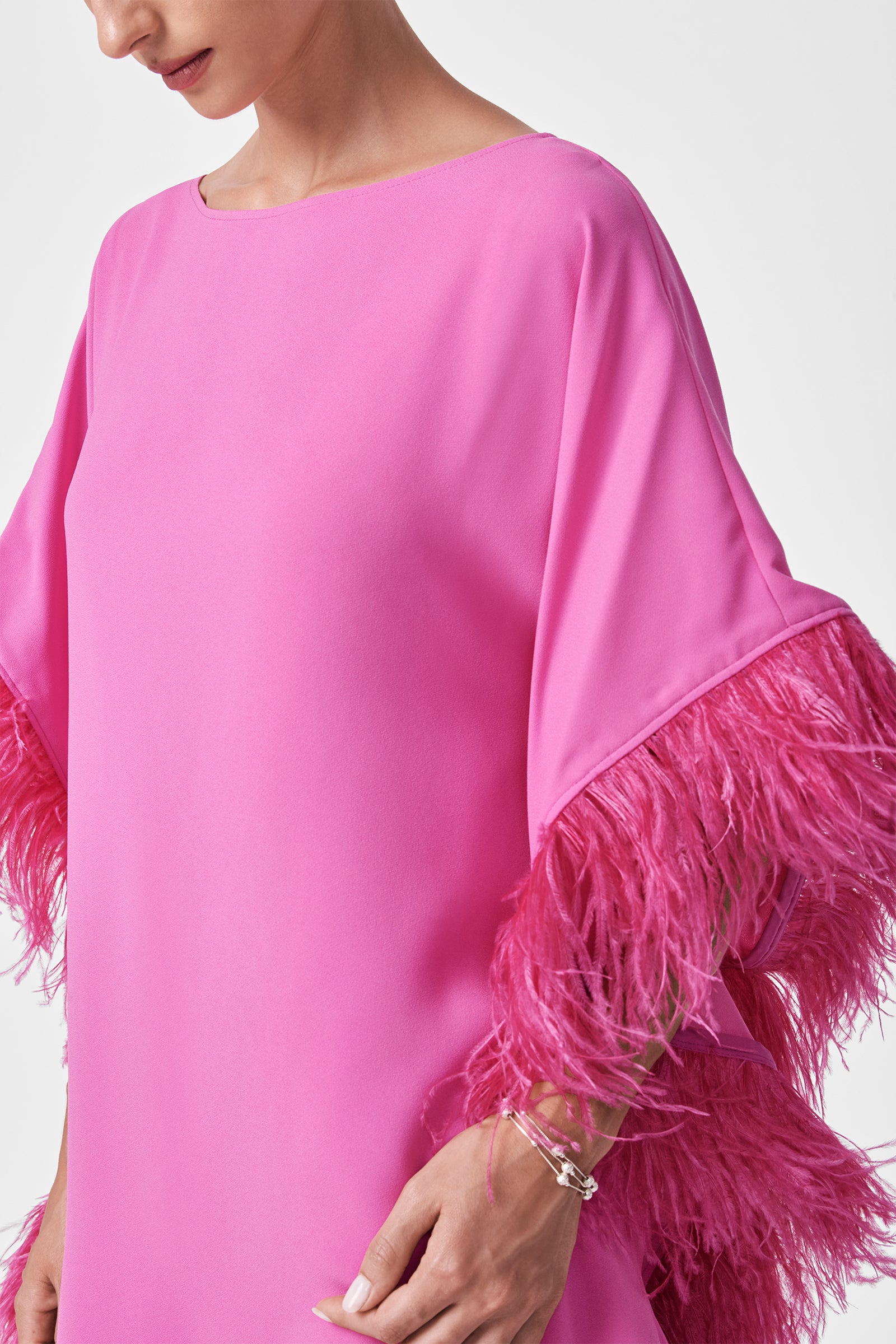 A woman wears the Mini Neena Caftan Dress—a loose, bright pink mini dress with wide sleeves and long dark pink feather trim—posing against a plain white background. Only her upper body and part of her face are visible.