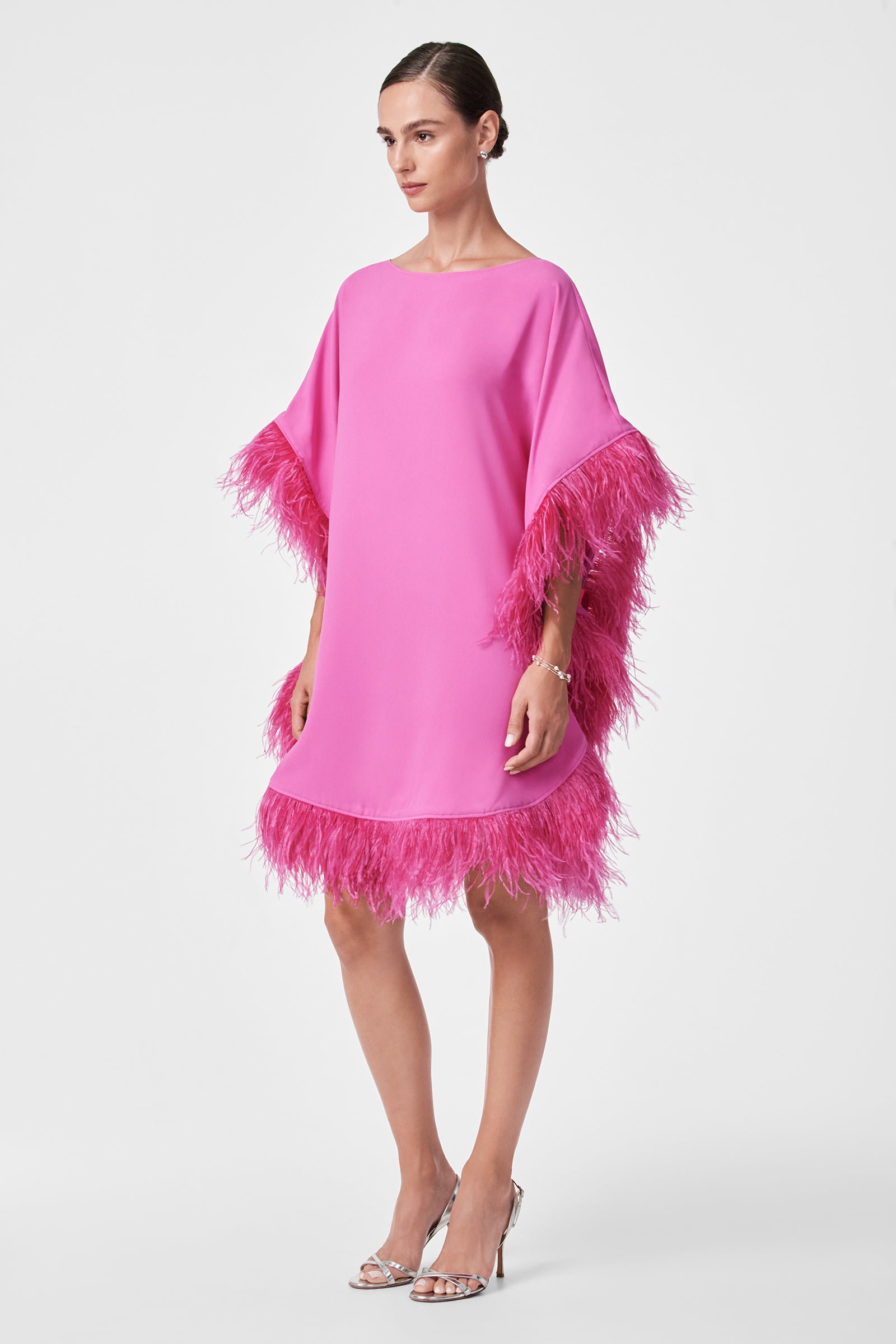 A woman poses against a plain white background wearing the Mini Neena Caftan Dress, a bright pink, loose-fitting piece with feather trim on the sleeves and hem. She completes the look with silver high-heeled sandals and has her hair pulled back.