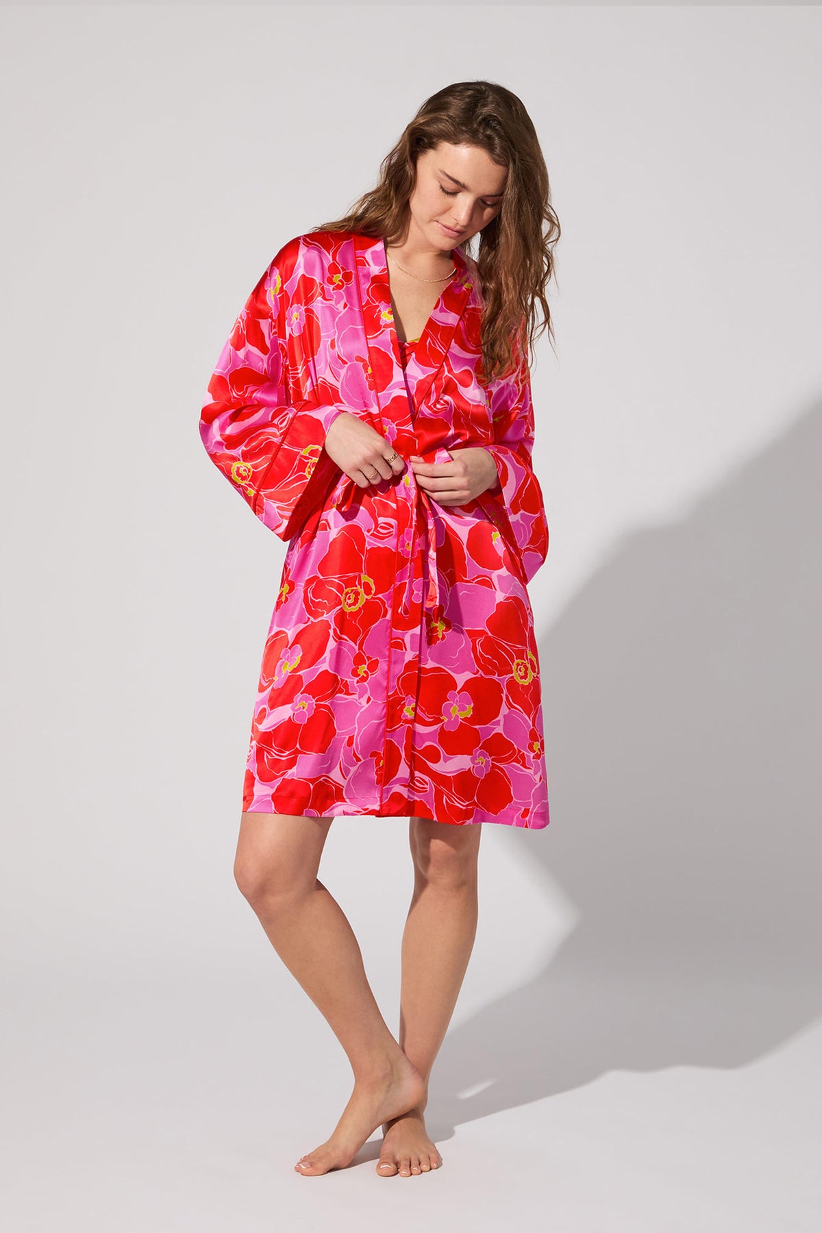 A woman stands barefoot against a plain background, tying the belt of the Long-Sleeve Silk Robe in Winter Orchids, which features wide sleeves and a vibrant floral design.
