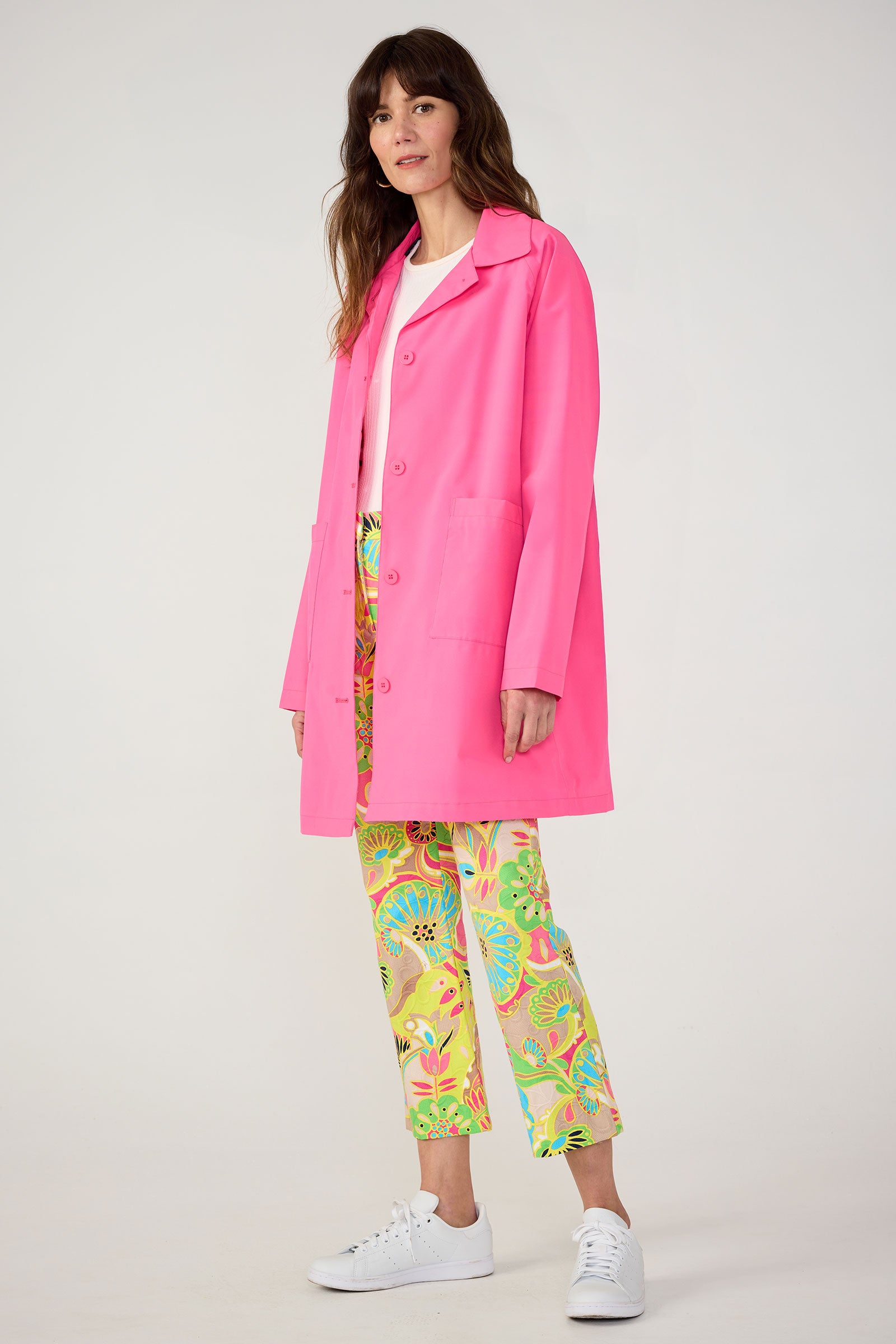 A woman with long brown hair poses against a light background wearing the TT X SR Mac Rain Coat in pink over a white shirt and colorful patterned pants with floral and animal designs, paired with white sneakers.