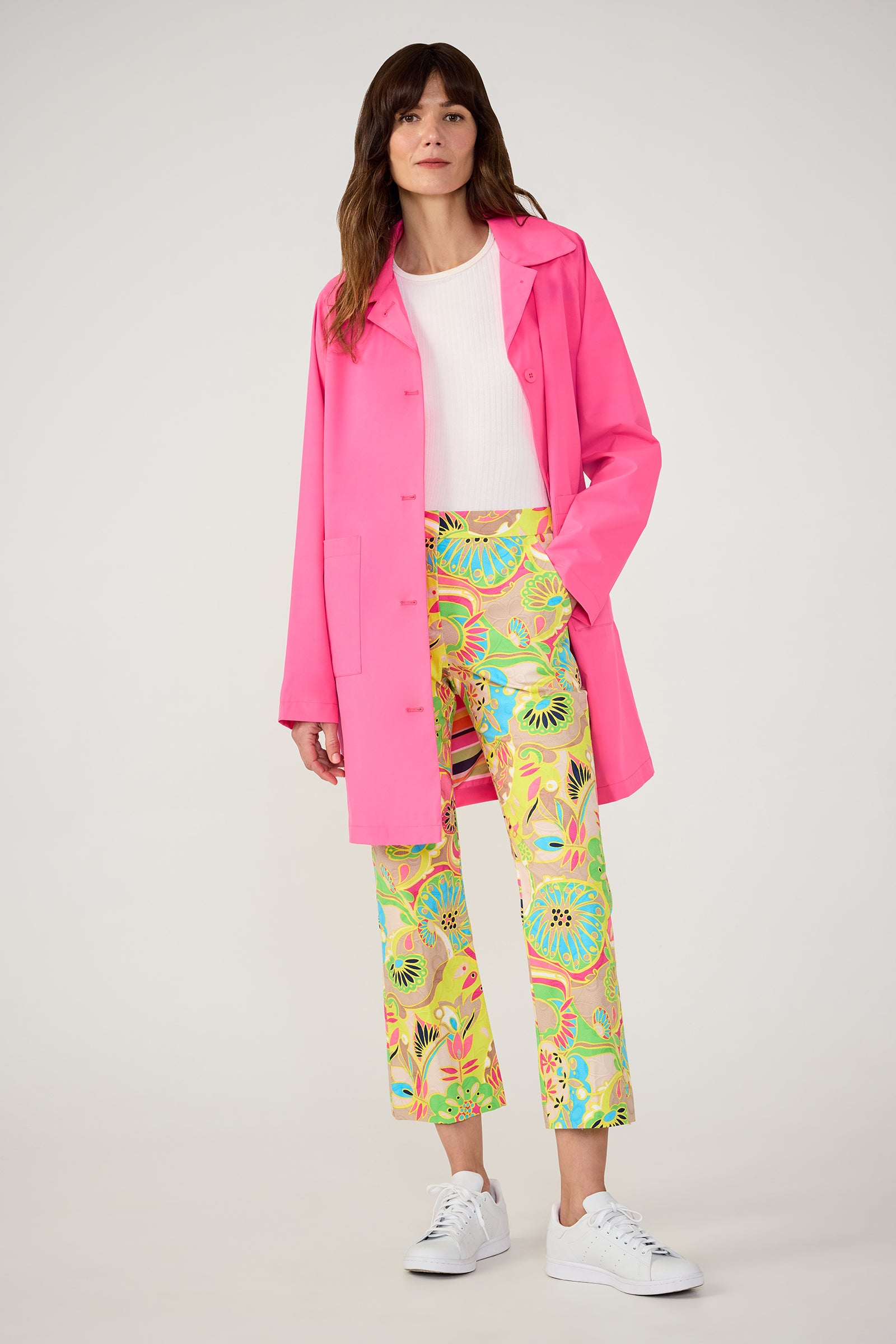 A woman stands wearing the TT X SR Mac Rain Coat in pink over a white top, floral print pants, and white sneakers against a plain light background.
