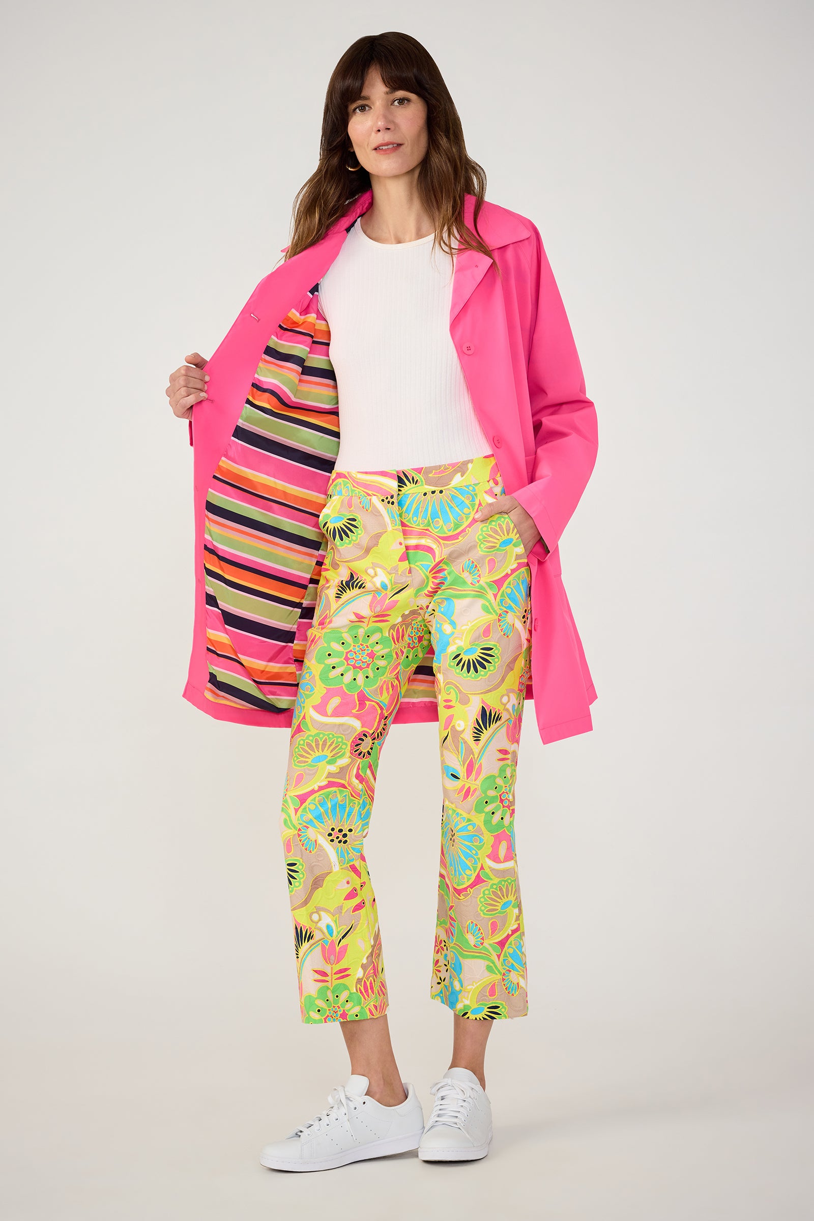 A woman wears the TT X SR Mac Rain Coat in pink, featuring a colorful striped lining, over a white ribbed top, floral print pants, and white sneakers against a plain background.