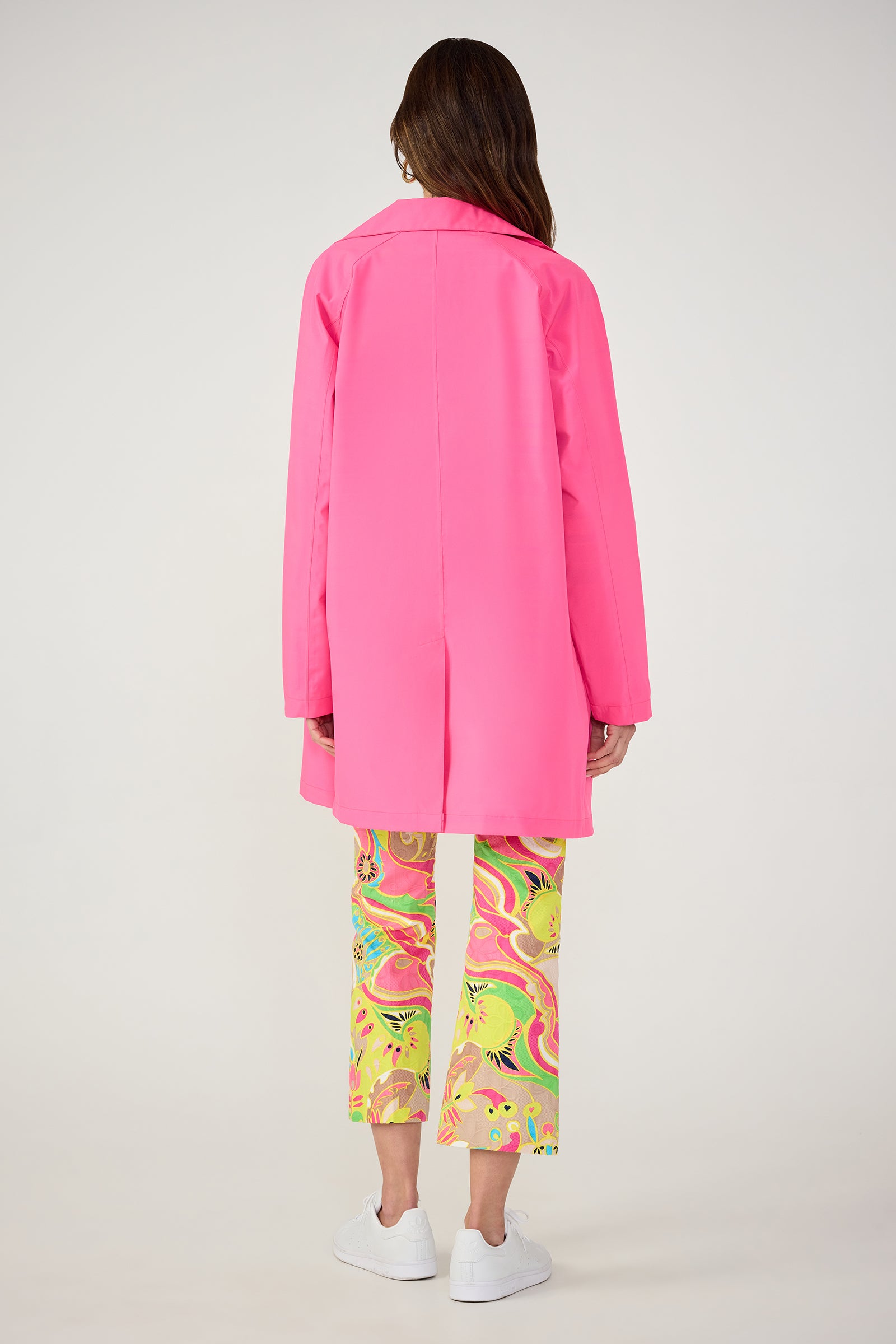 A person faces away, wearing the TT X SR Mac Rain Coat in pink, paired with colorful floral pants and white sneakers, set against a plain light background.