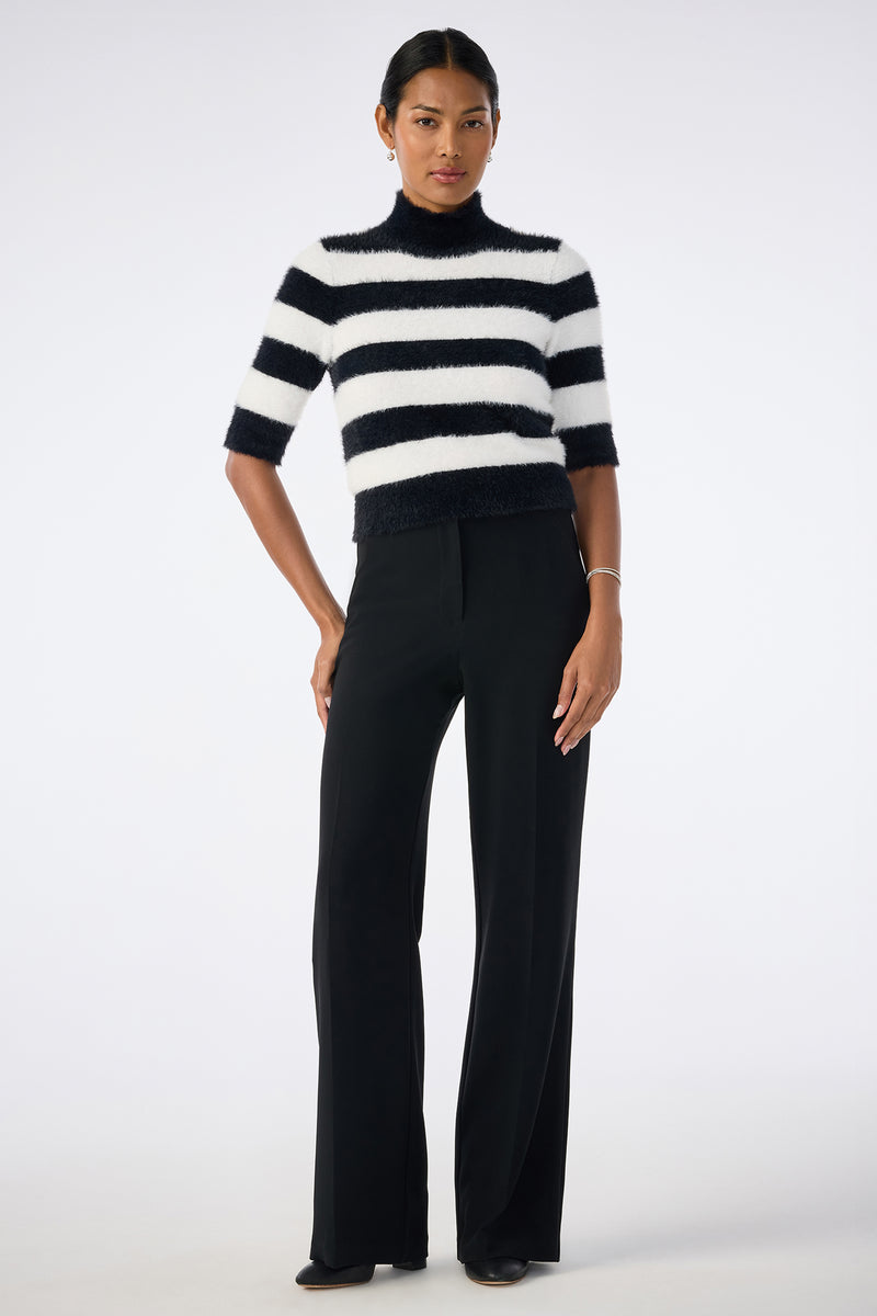 A woman stands facing forward against a plain light background, wearing the Mali Mock Neck Sweater in black and white stripes, paired with black wide-leg trousers and dark shoes. Her hair is pulled back.