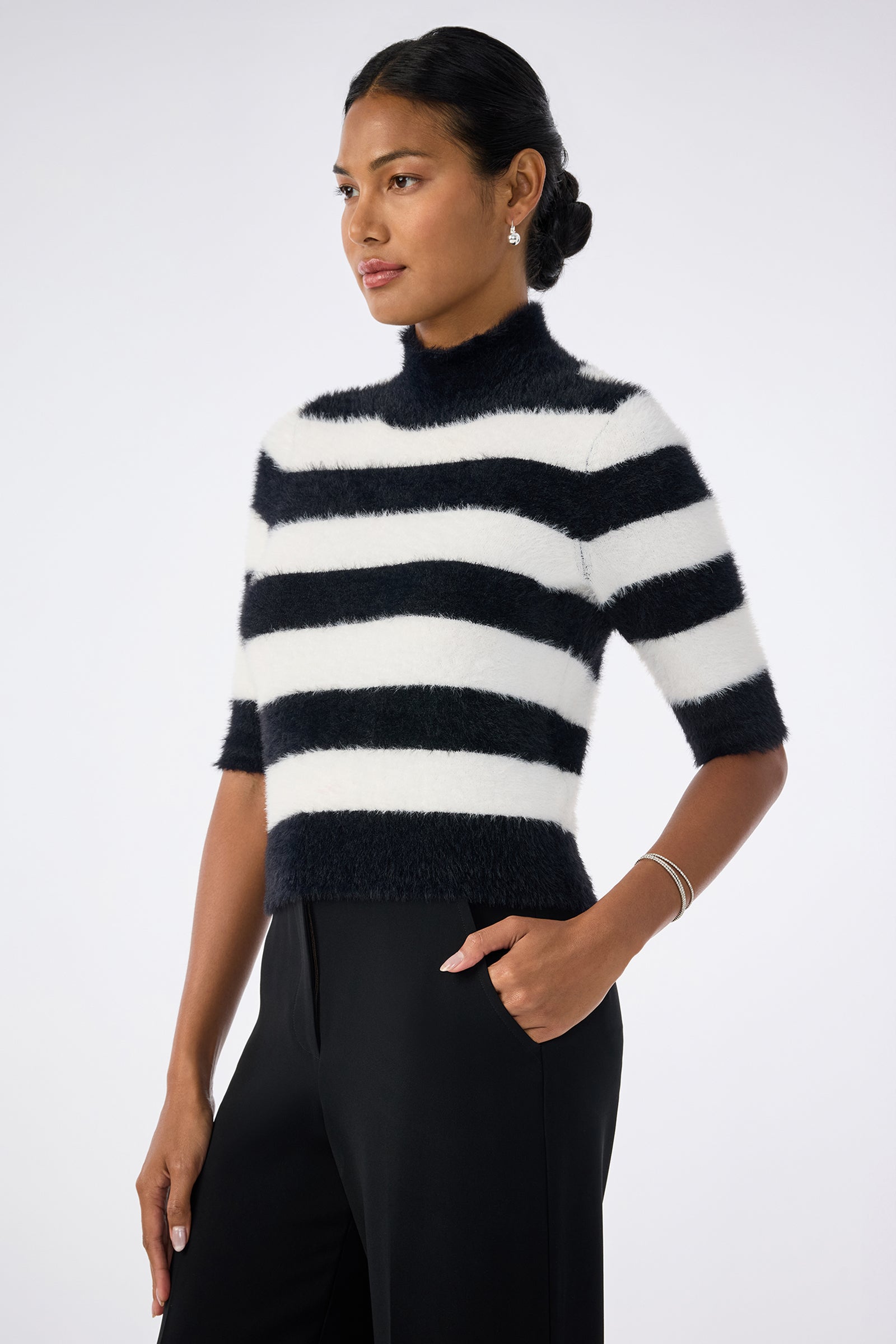 A woman with dark hair in a low bun wears the Mali Mock Neck Sweater—fuzzy, short-sleeved, black and white striped—paired with black pants, standing against a plain light background with one hand in her pocket.