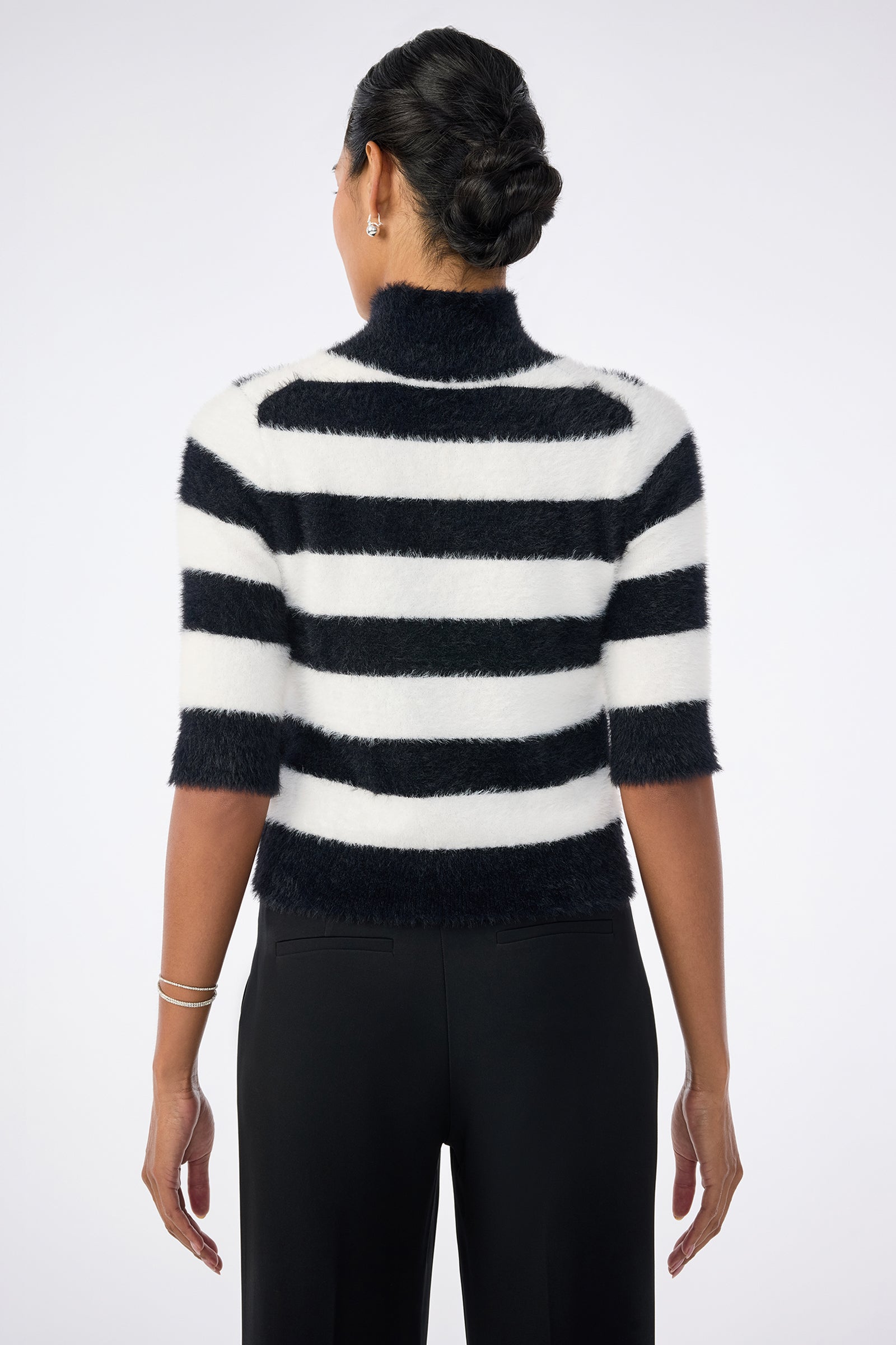 A person with dark hair in a braided updo is seen from behind, wearing the Mali Mock Neck Sweater, a fuzzy black and white horizontally striped short-sleeve style, paired with black pants against a plain light background.