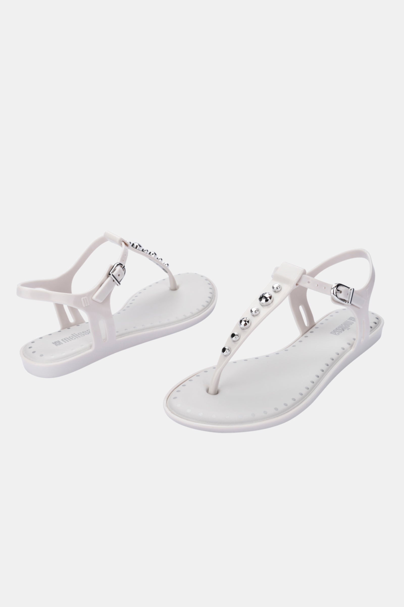 Melissa Solar Boho: A pair of white flat sandals featuring ankle straps, metallic studs on the T-strap, and silver buckles, showcased against a plain white background.