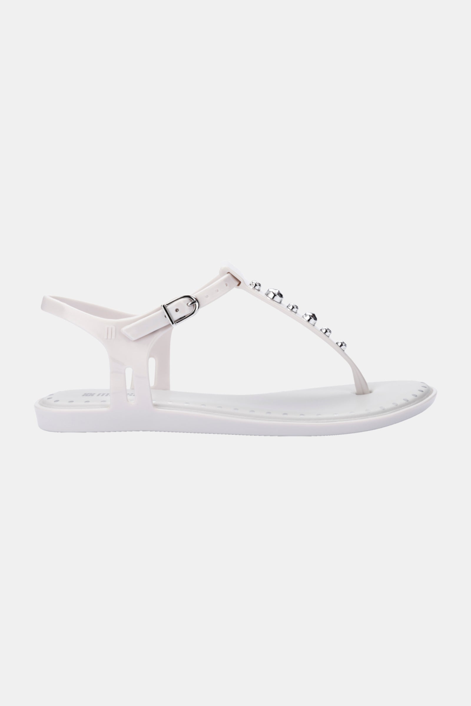 The Melissa Solar Boho is a white flat sandal featuring a T-strap design with silver studs and a small adjustable ankle buckle, displayed on a plain light background.