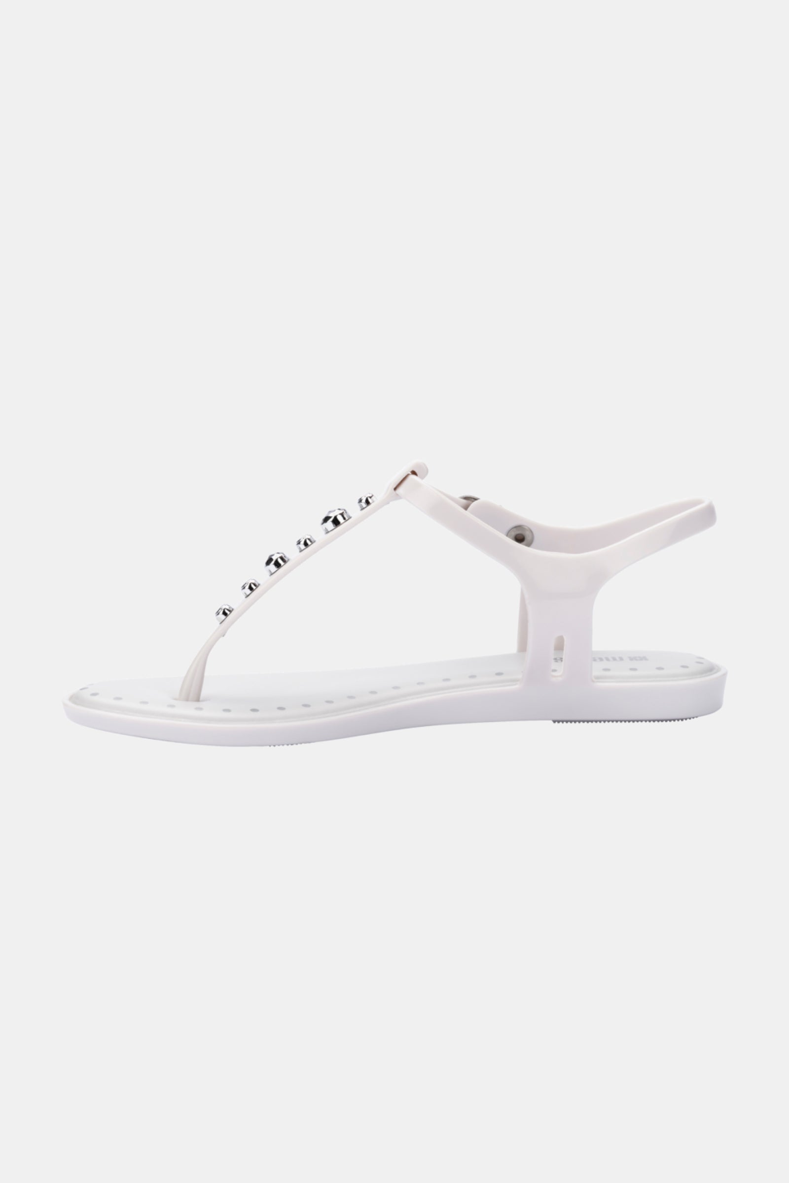 The Melissa Solar Boho is a white flat sandal with a minimalist thong design, featuring a T-strap embellished with round metallic studs and an ankle strap with button closure, showcased on a plain background.