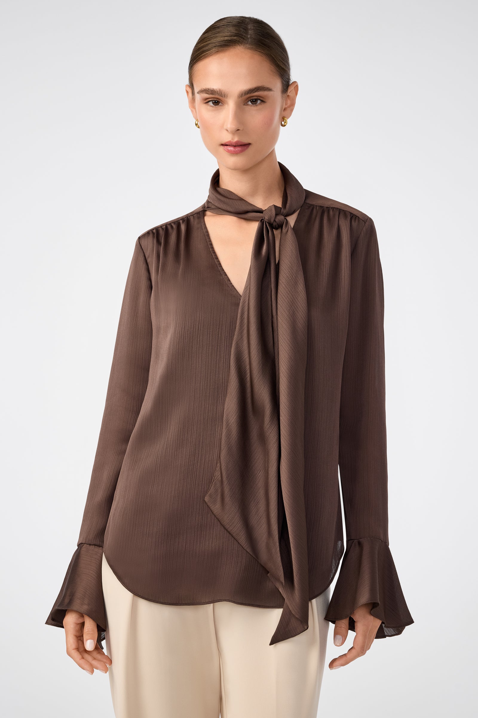 A woman wears the Alora Lavalliere Blouse in walnut, featuring long sleeves, a tie-neck detail, and flared cuffs, paired with light-colored pants, standing against a plain, light background.