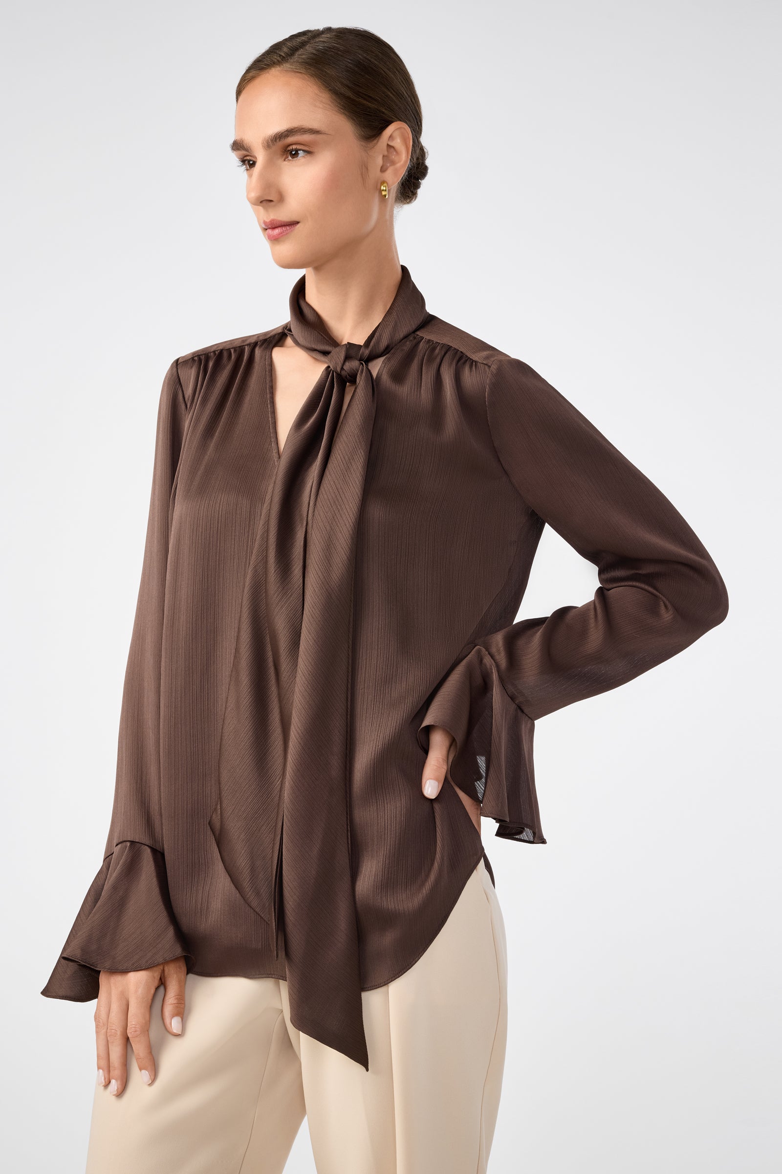 A woman stands against a plain background, wearing the Alora Lavalliere Blouse—a silky brown long-sleeve top with a tie-neck and flared cuffs—paired with cream-colored pants, one hand on her hip.