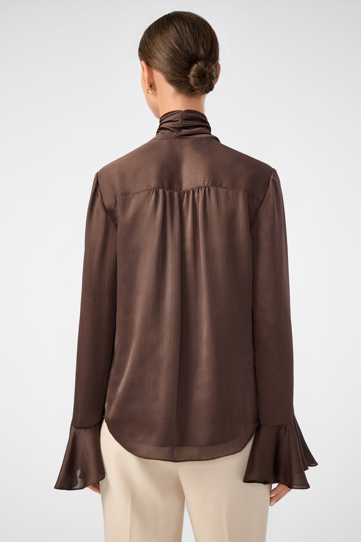 A woman with her hair in a low bun is shown from the back, wearing the Alora Lavalliere Blouse—long-sleeved and dark brown with flared cuffs—paired with cream-colored pants against a plain light background.