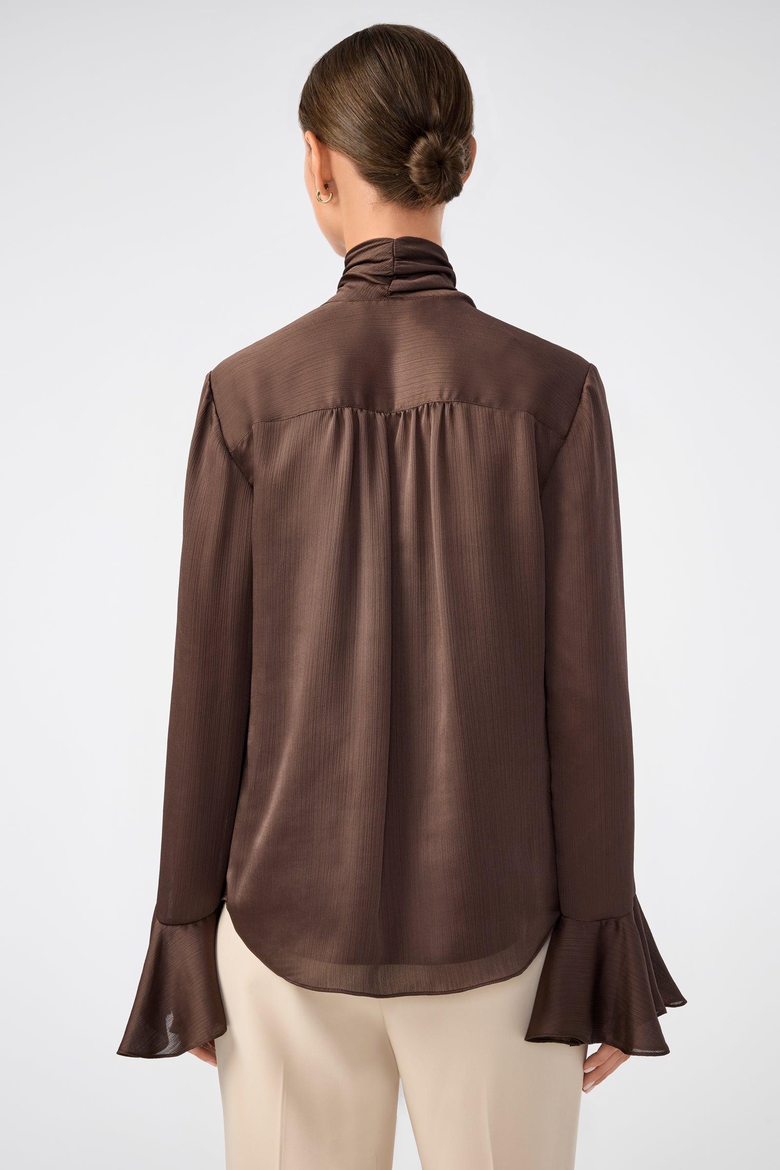 A woman with her hair in a low bun is shown from the back, wearing the Alora Lavalliere Blouse—long-sleeved and dark brown with flared cuffs—paired with cream-colored pants against a plain light background.