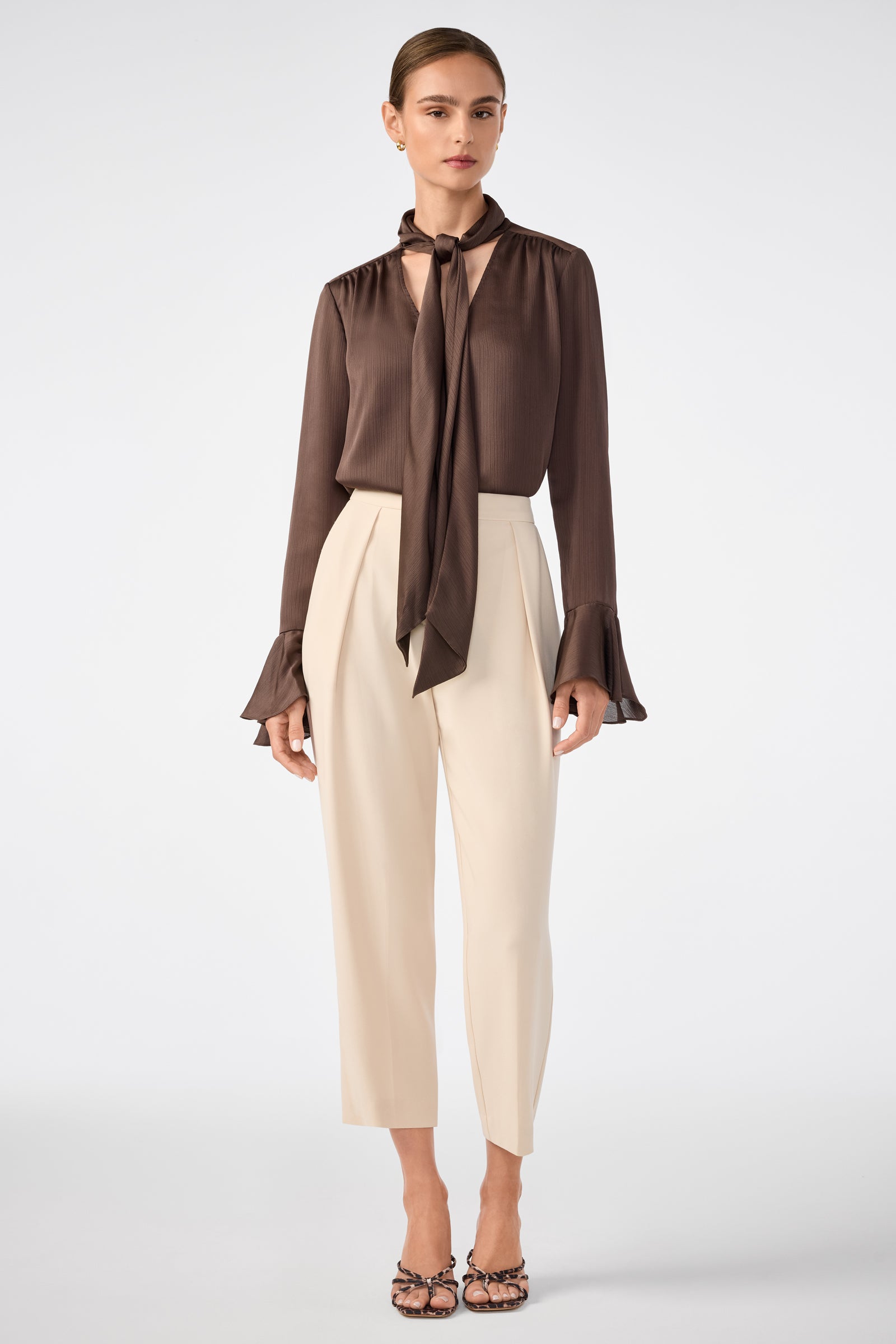 A woman wears the Alora Lavalliere Blouse, a brown long-sleeve top with a neck tie and flared cuffs, paired with high-waisted cropped cream trousers and heels, standing against a plain light background.