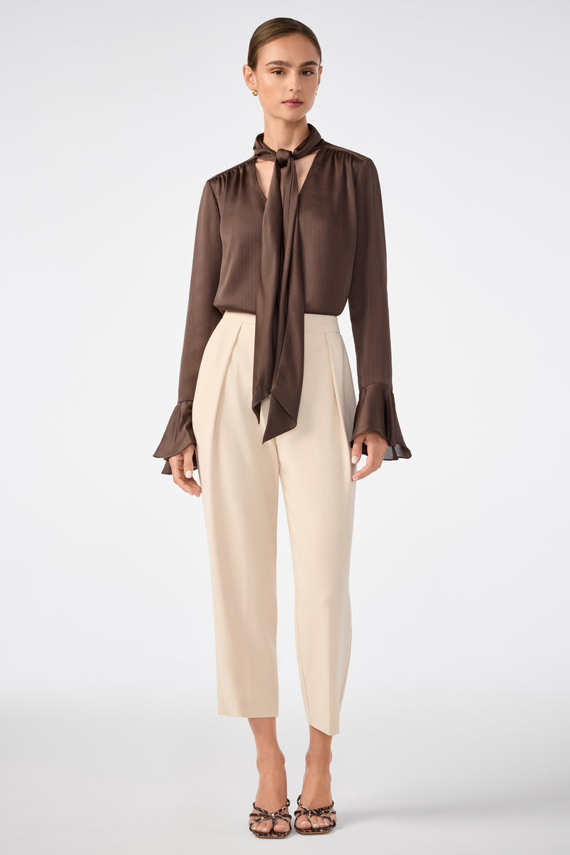 A woman wears the Alora Lavalliere Blouse, a brown long-sleeve top with a neck tie and flared cuffs, paired with high-waisted cropped cream trousers and heels, standing against a plain light background.