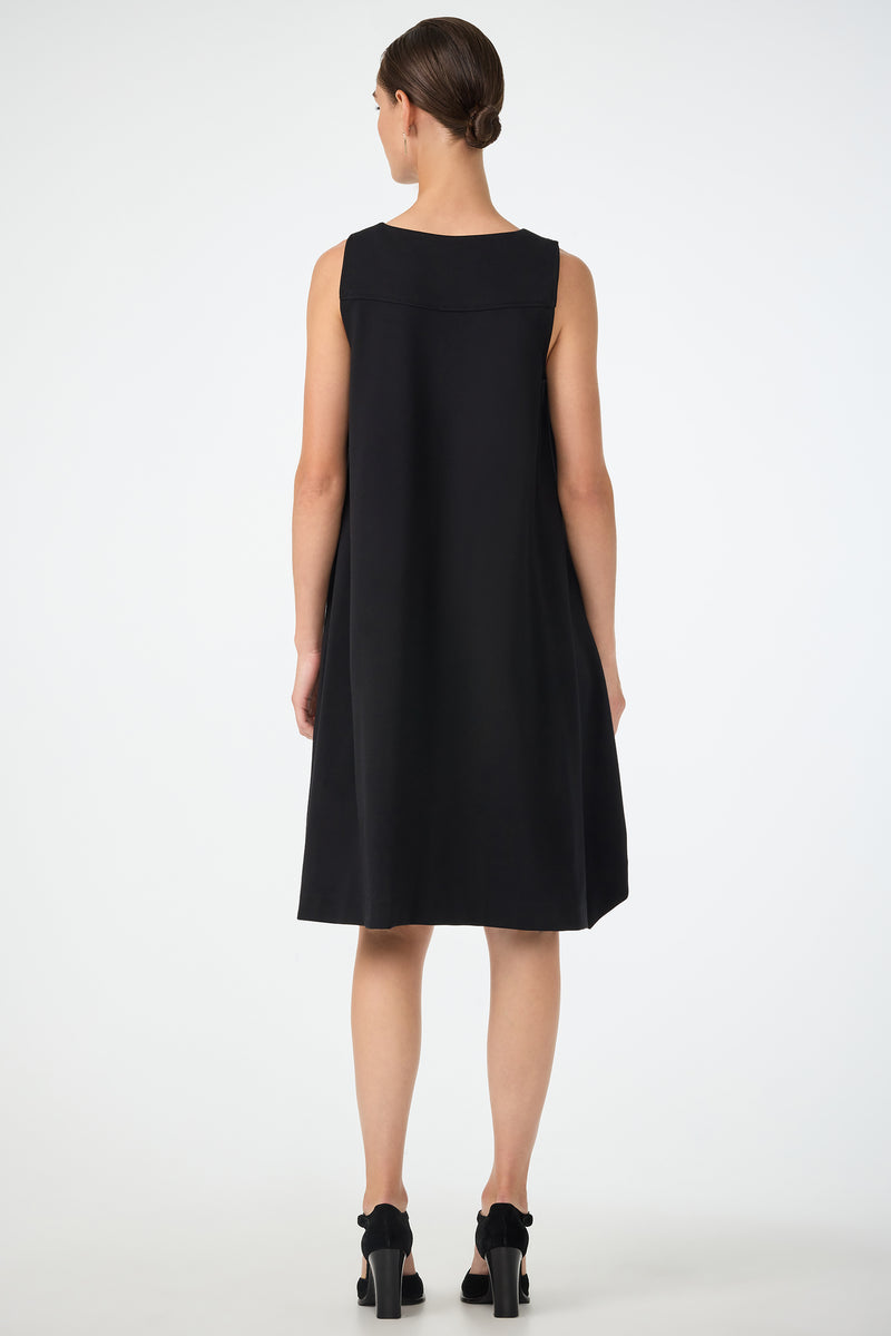 A woman faces away, wearing the Vantis High-Low Sleeveless Dress in black with black heels. Her hair is neatly tied back, and she stands against a plain white background.