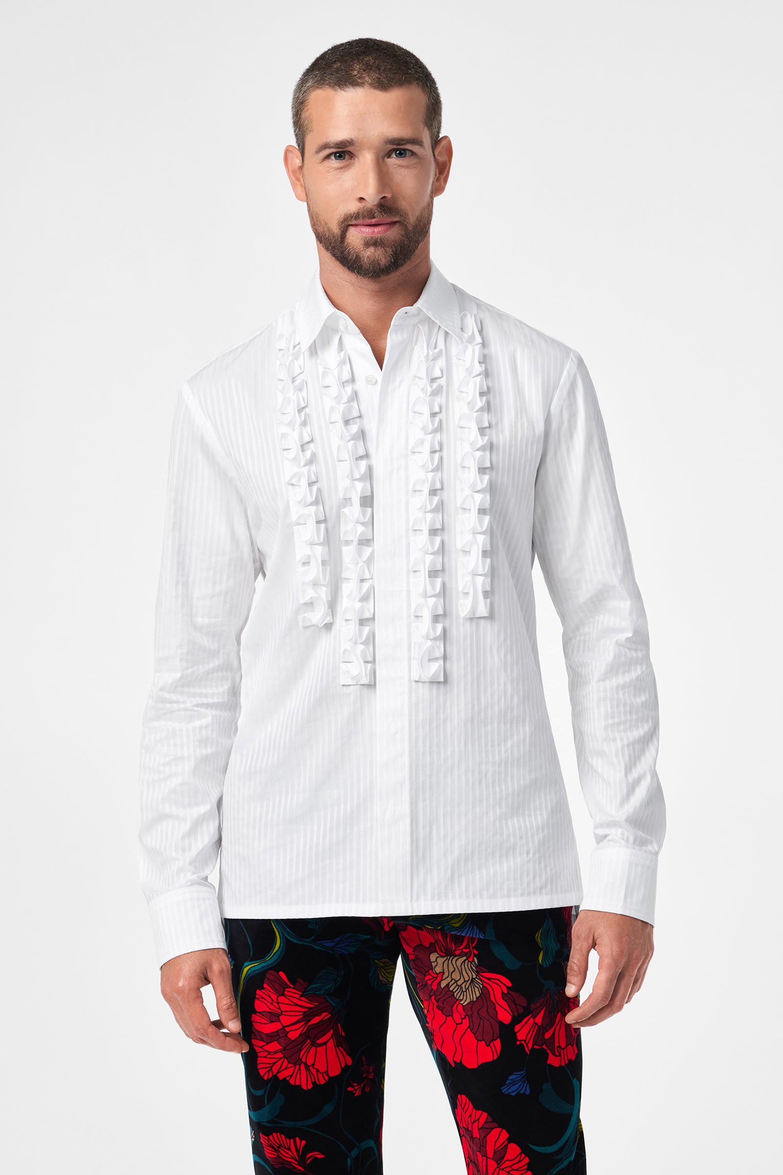 A man wears the Evan Tux Shirt, a white long-sleeve button-down with ruffled front detailing, paired with black pants featuring a bold red and blue floral pattern, standing against a plain white background.