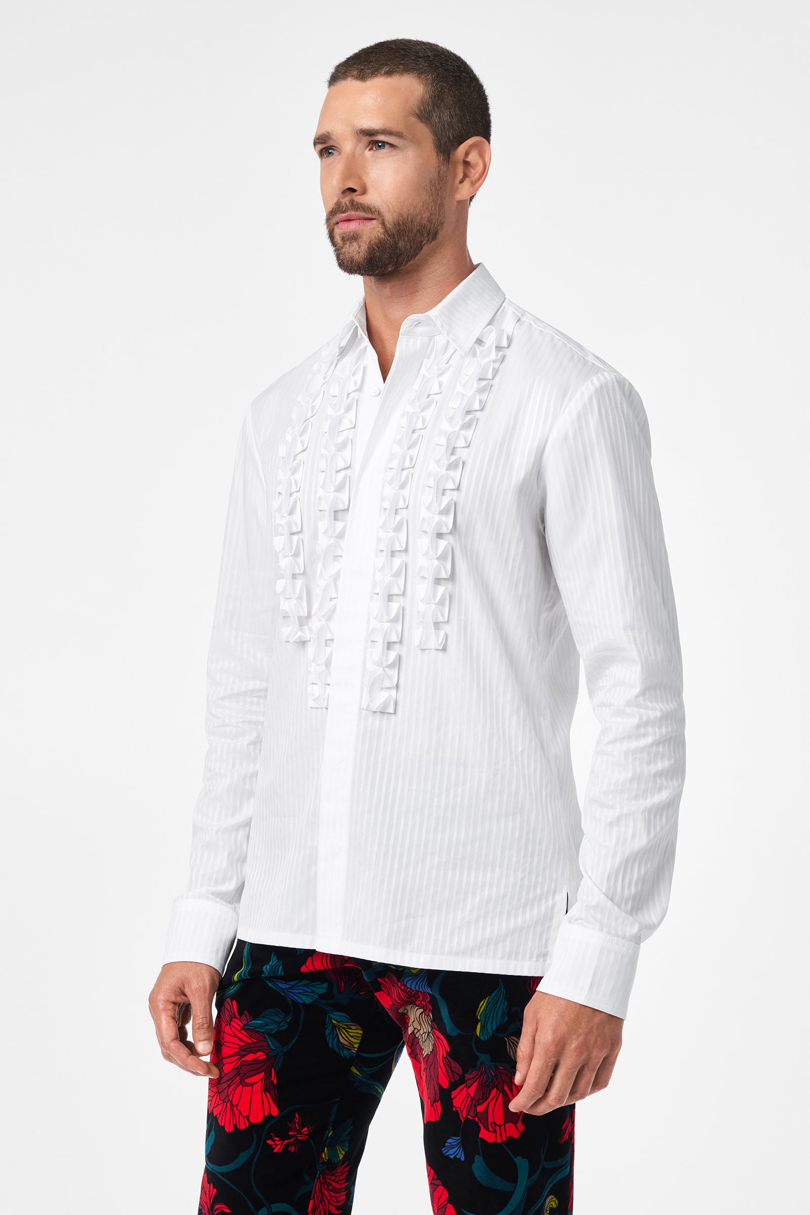 A man wears the Evan Tux Shirt, a white long-sleeve button-down with front ruffle details, paired with bold black pants featuring red floral prints, standing against a plain white background.