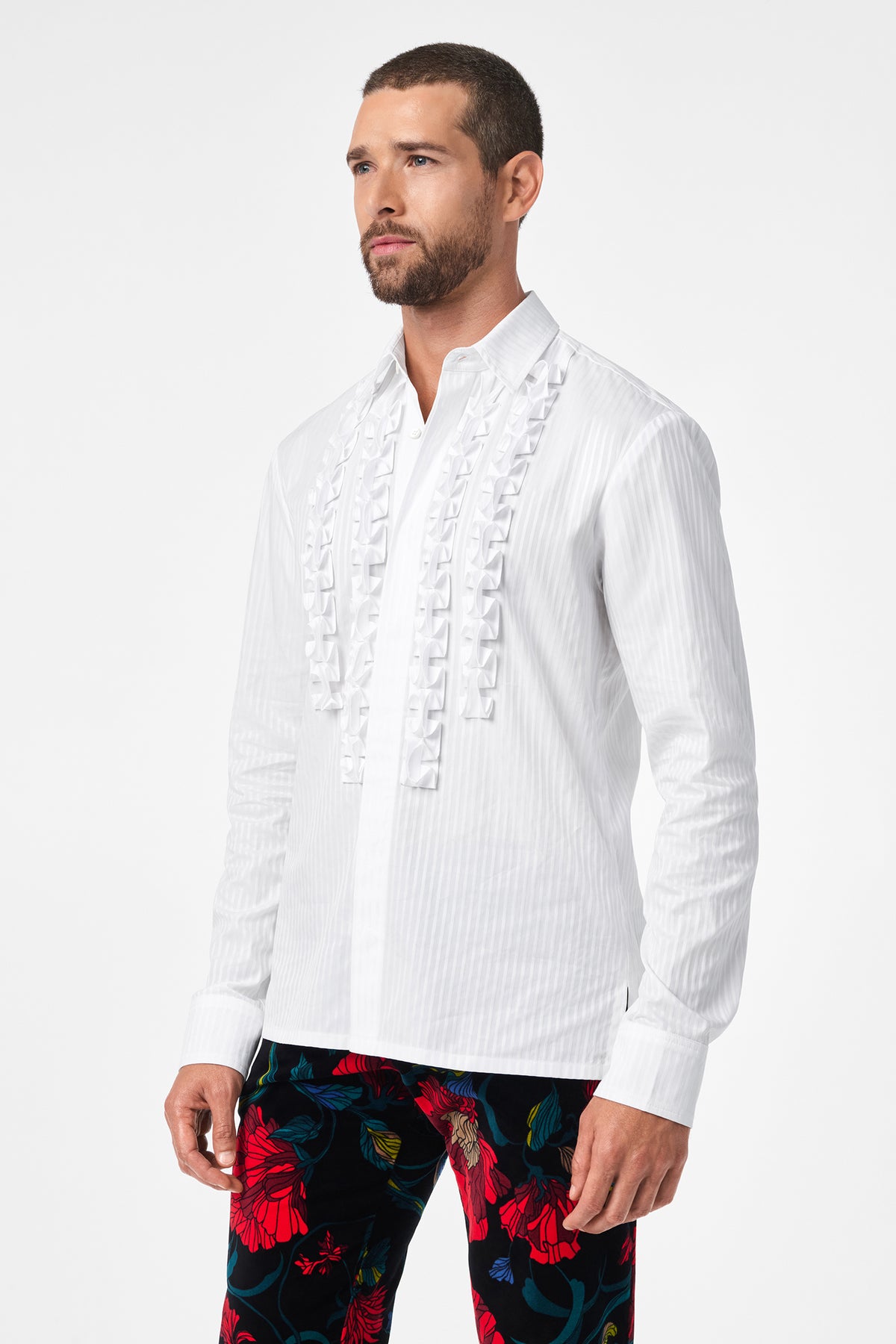 A man wears the Evan Tux Shirt, a white long-sleeve button-down with front ruffle details, paired with bold black pants featuring red floral prints, standing against a plain white background.
