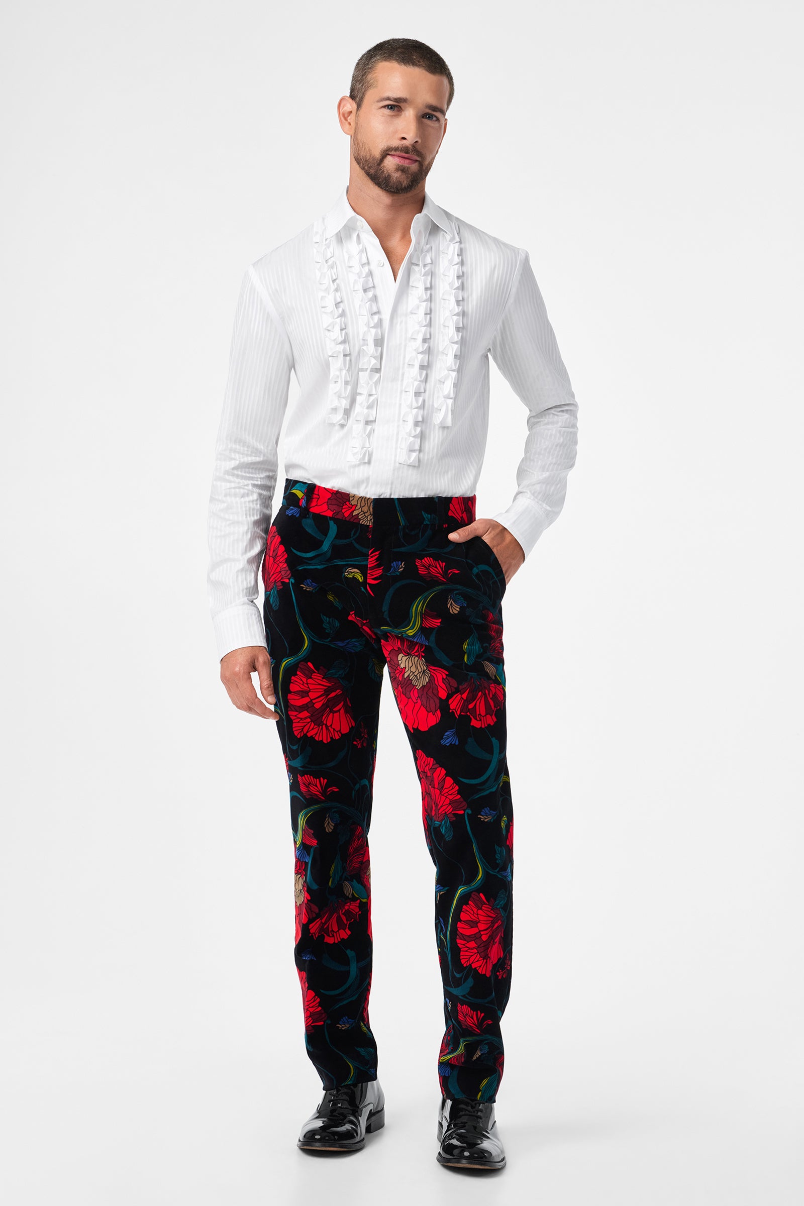 A man wears the Evan Tux Shirt, a white ruffled dress shirt, with black velvet pants featuring a bold red floral pattern and black shoes, standing against a plain white background.