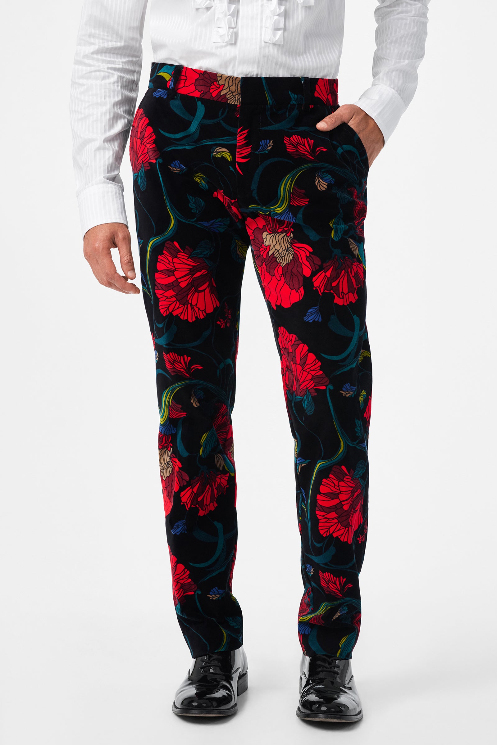 A person wearing the Clyde Slim Printed Velvet Trouser, featuring a bold red and green floral pattern, paired with a white ruffled long-sleeve shirt and shiny black dress shoes.