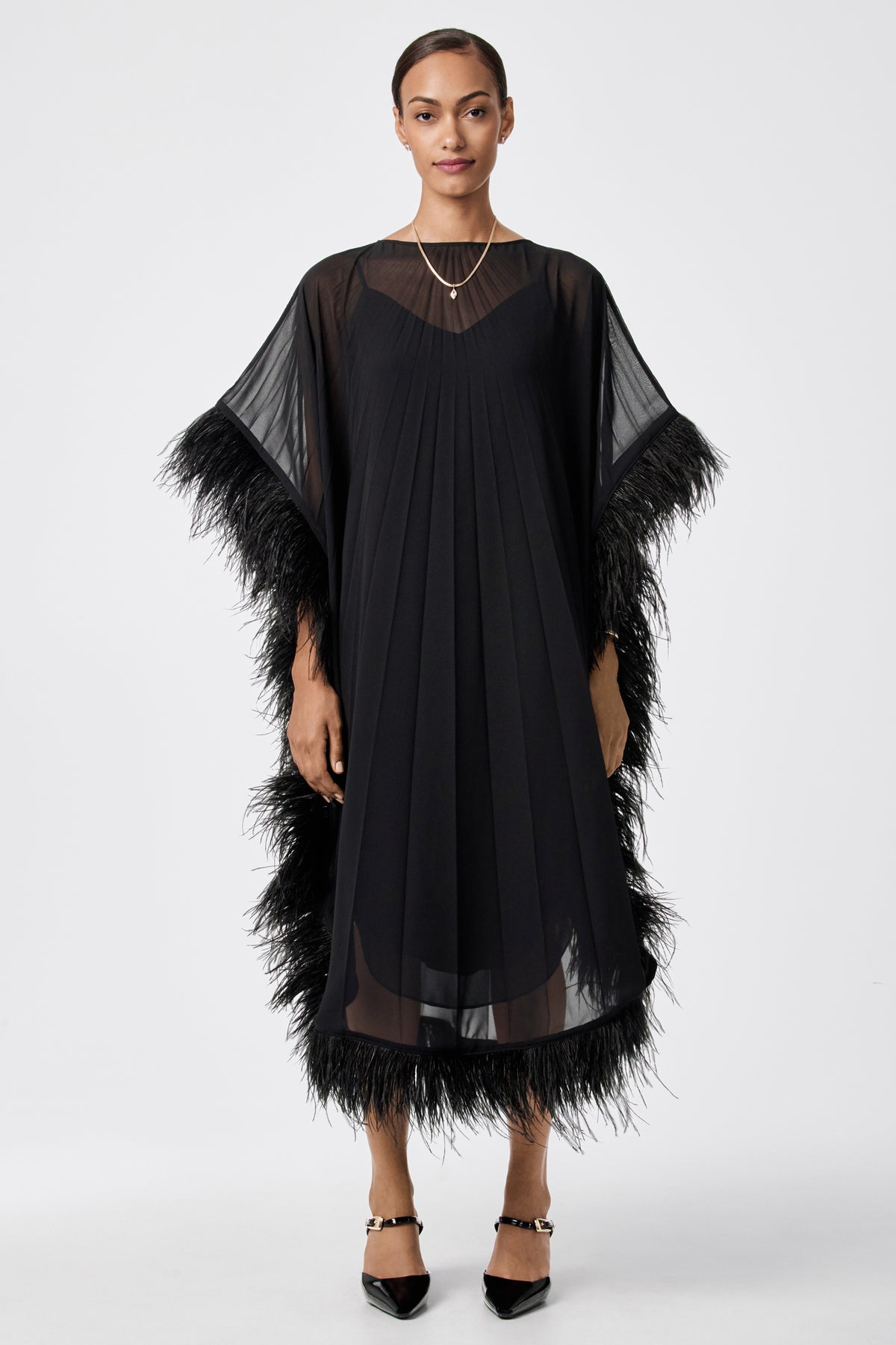 A woman stands facing forward in the Pleated Neena Feather-Trimmed Caftan Dress, featuring sheer sleeves and feather trim. She has short hair, minimal jewelry, and wears black pointed shoes against a plain white background.