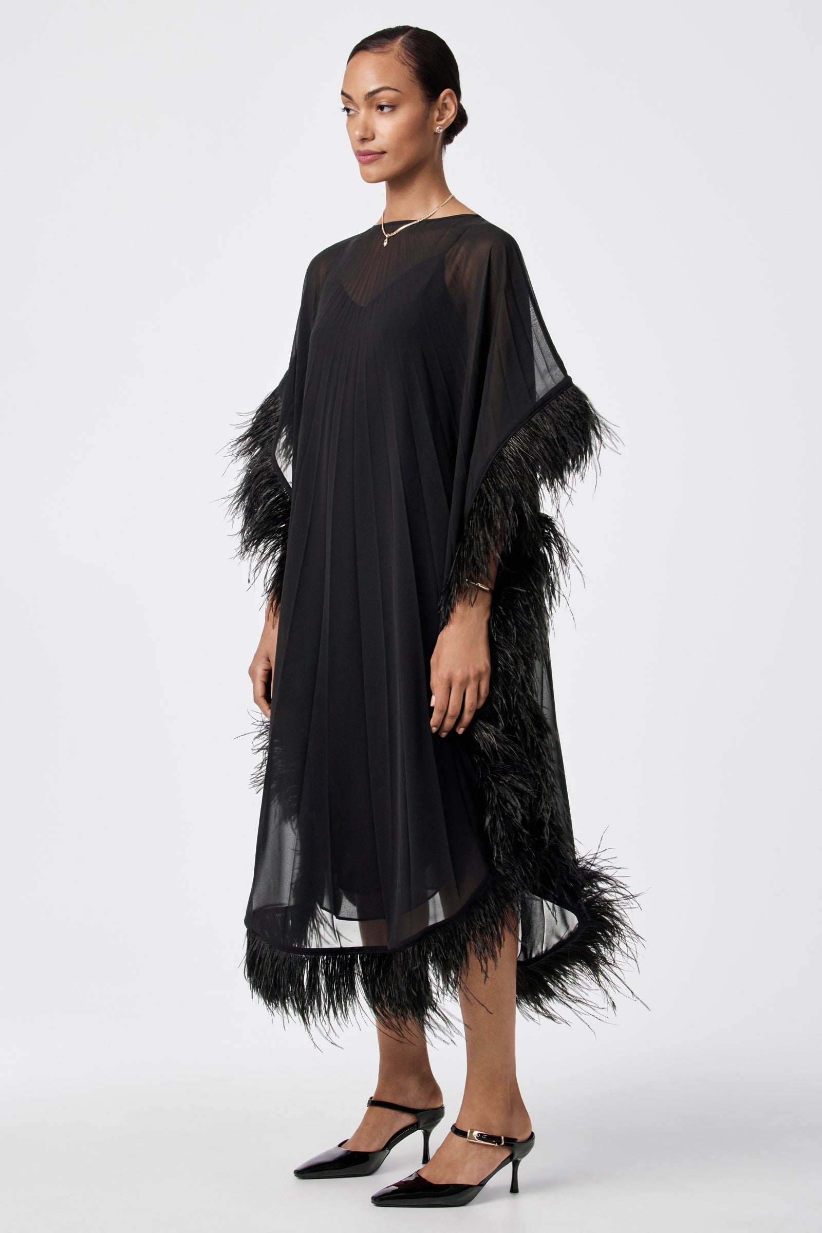 A woman stands against a plain white background wearing the Pleated Neena Feather-Trimmed Caftan Dress, featuring sheer black fabric with feather trim at the sleeves and hem, paired with black heels and a simple necklace.