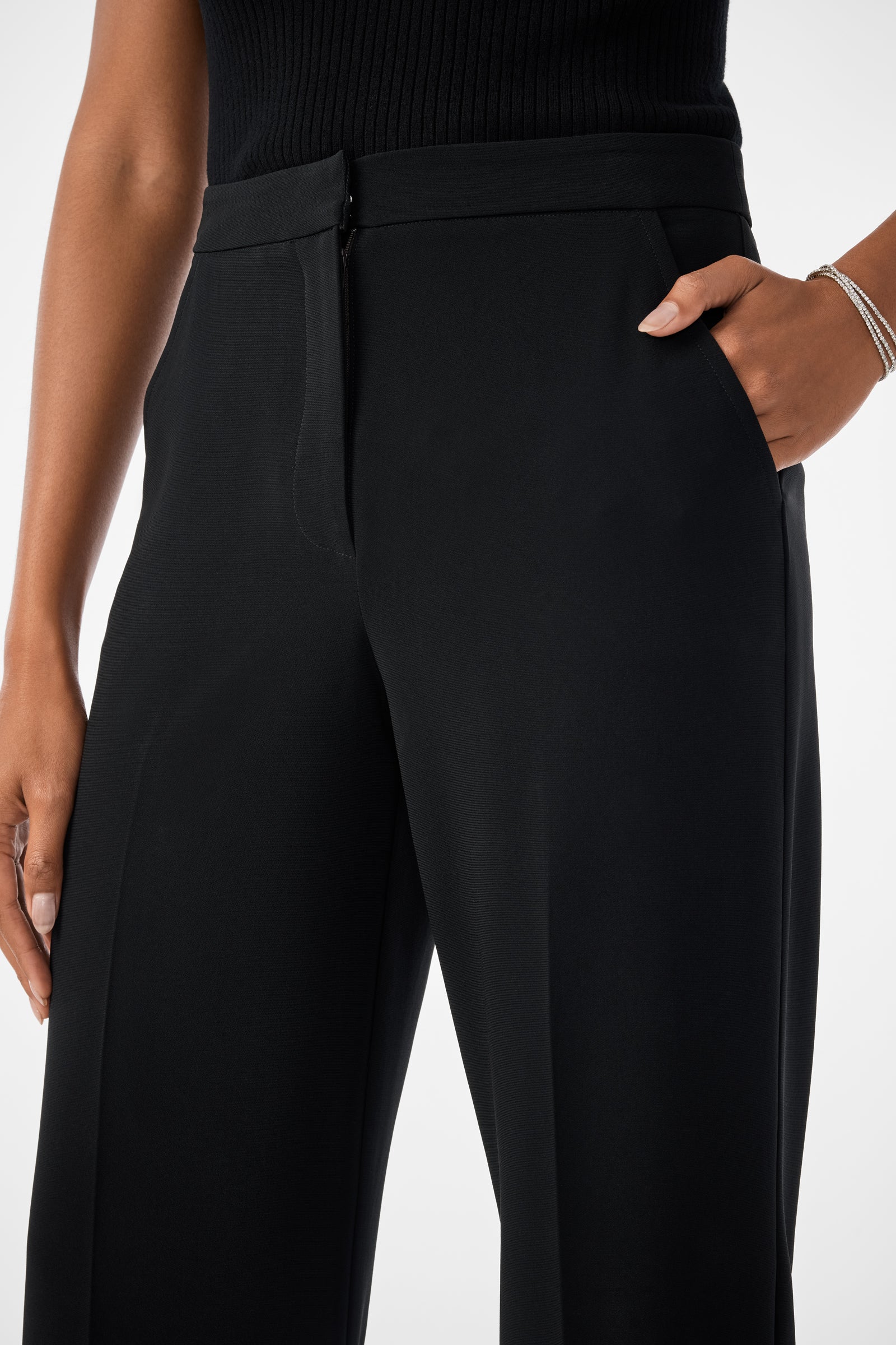 A person wears Sadie Wide-Leg Pants in black, featuring a high waist, side zipper, wide legs, and pockets. Theyre shown from waist to mid-thigh with one hand in a pocket, styled with a sleeveless black top and a bracelet.