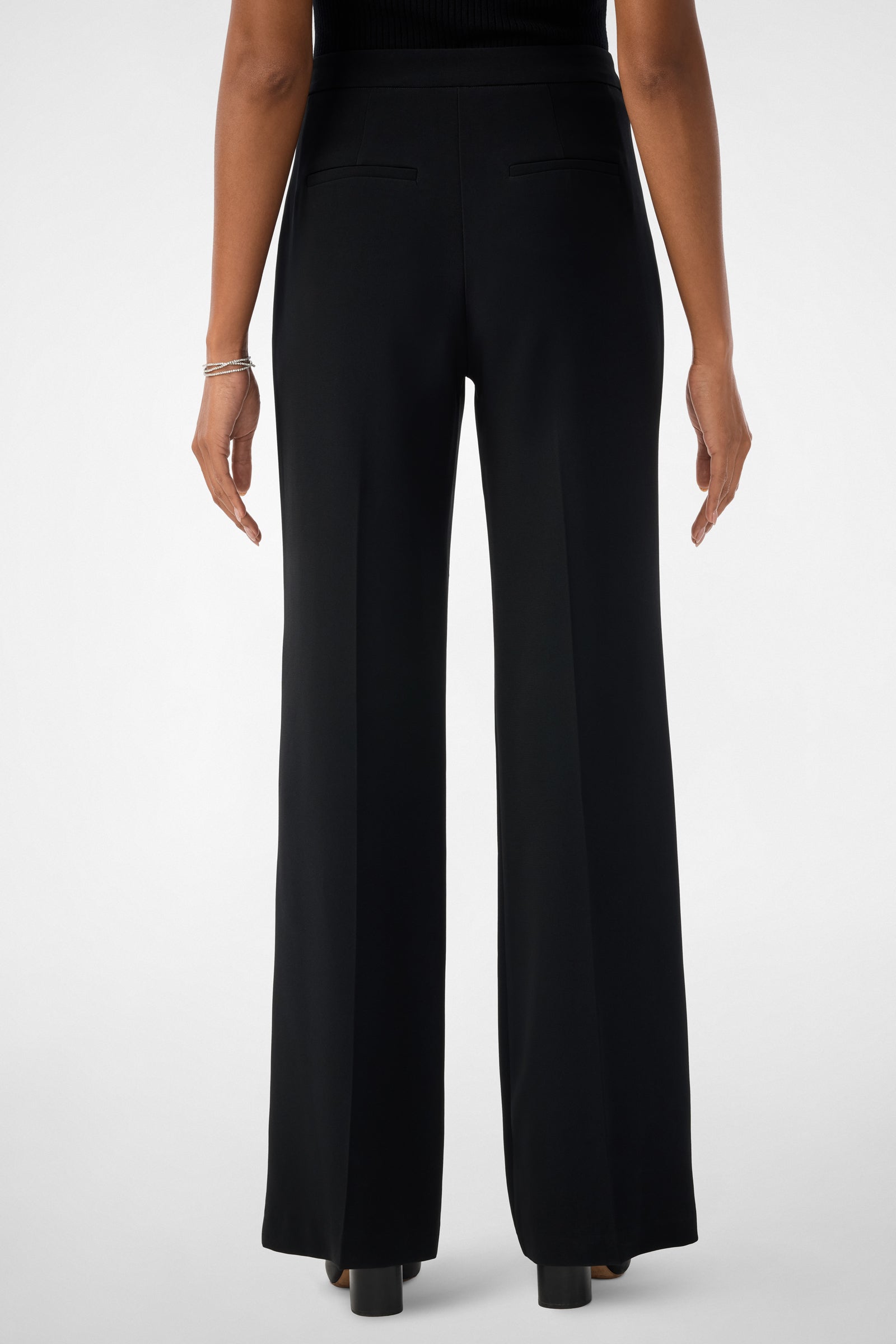 A person is shown from the back wearing the Sadie Wide-Leg Pants in black, featuring two back welt pockets, paired with a sleeveless black top, black heeled shoes, and a silver bracelet.