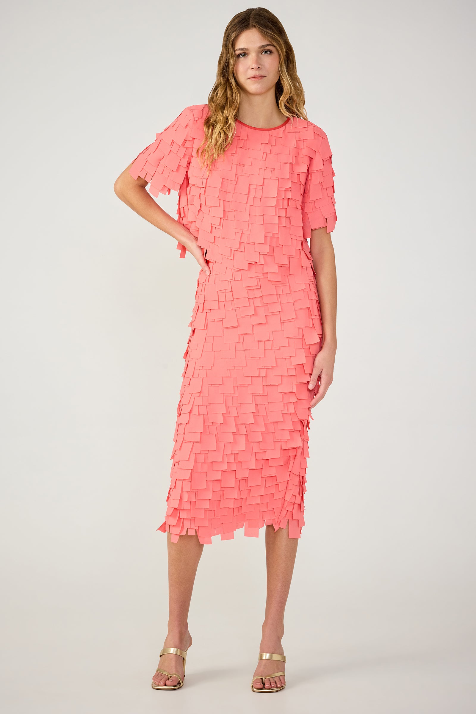 A woman wears the Dita Midi Pencil Skirt in Flutter Chiffon, a knee-length coral pink skirt with layered square details, styled with gold strappy sandals. She has long wavy hair and a neutral expression.