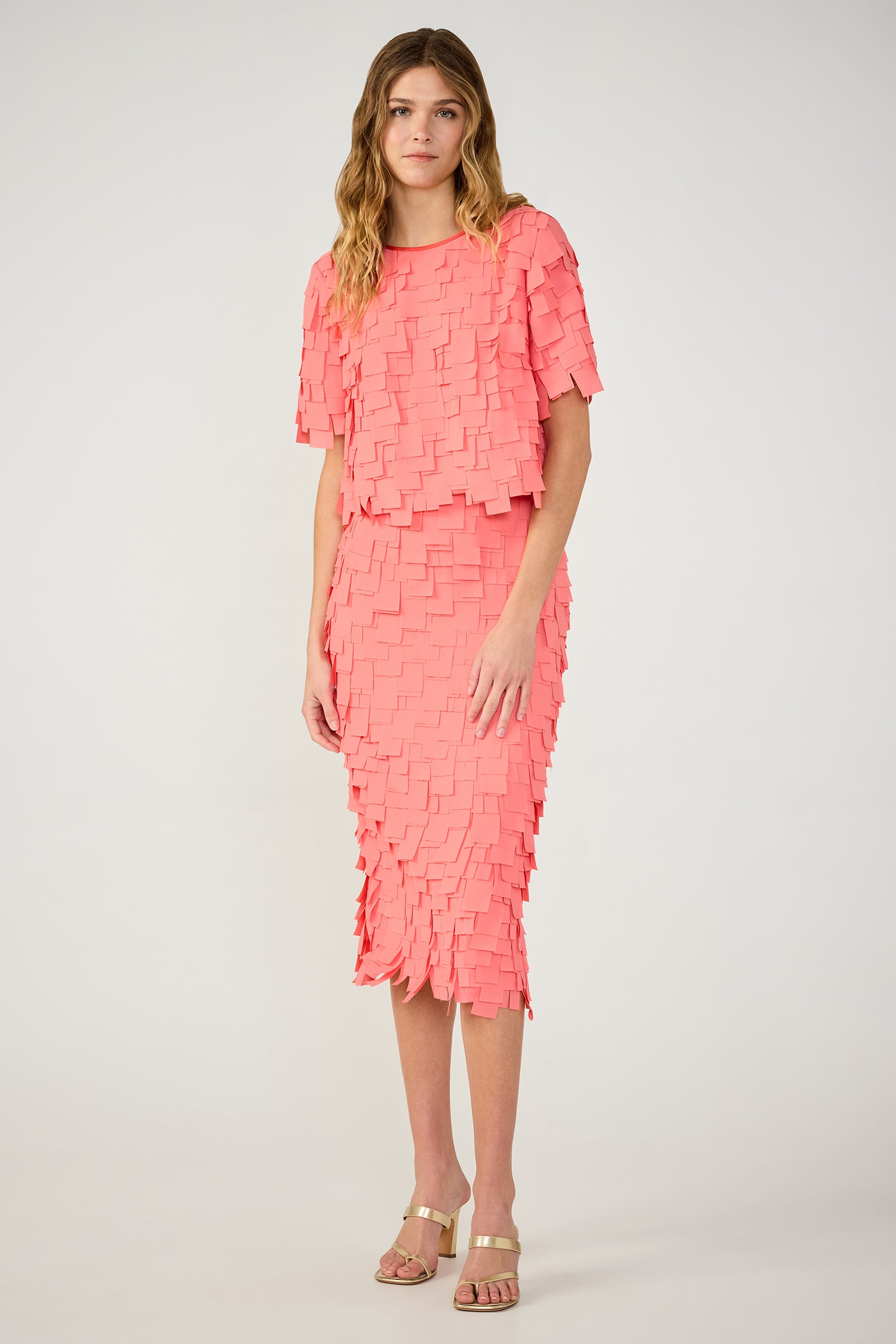 A woman stands against a plain background wearing the Serilda Short-Sleeve Top - Flutter Chiffon in coral pink, featuring textured overlapping square fabric pieces and paired with nude strappy sandals.