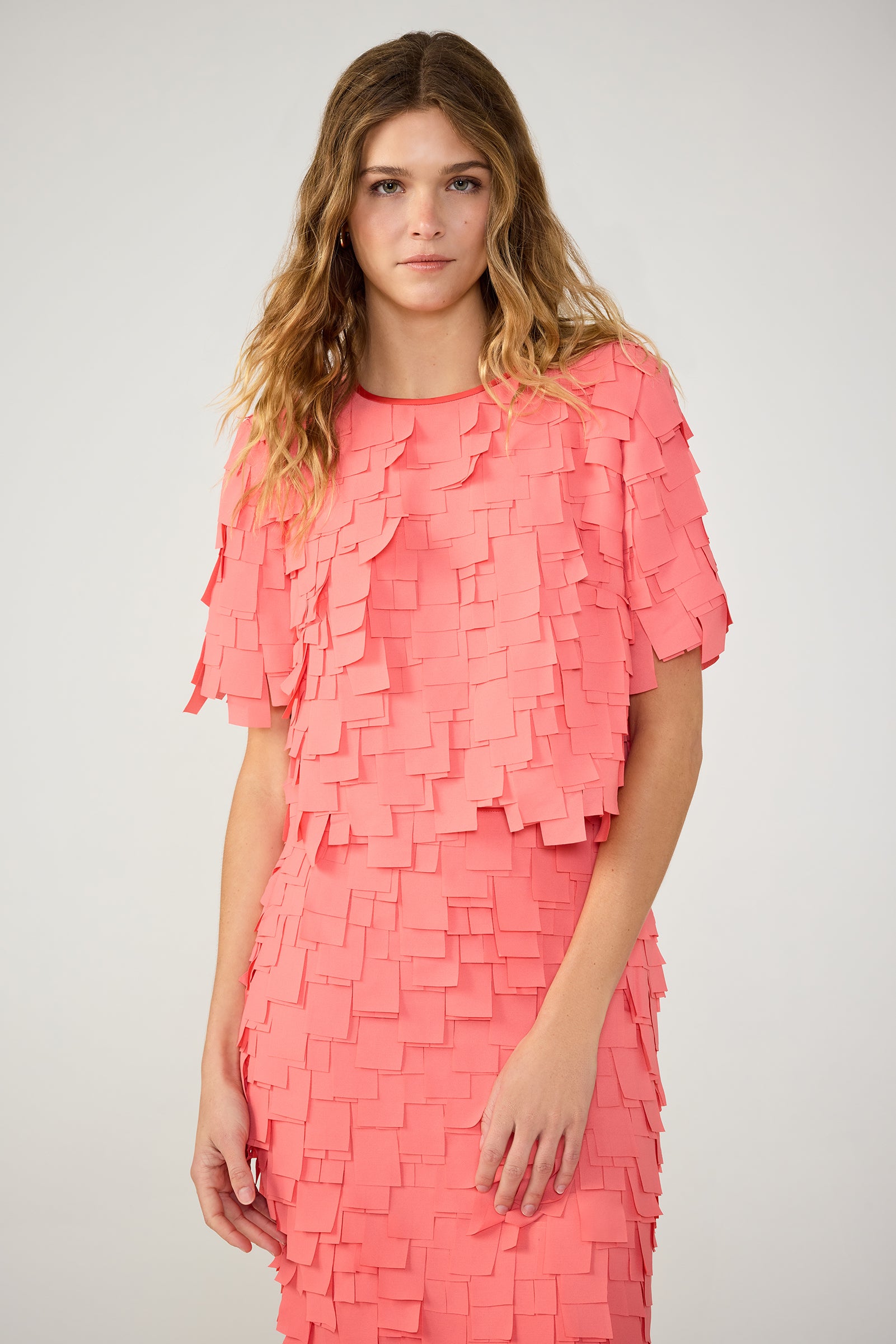 A woman with wavy, light brown hair wears the Serilda Short-Sleeve Top – Flutter Chiffon in Coral Crush, standing against a plain light gray background.