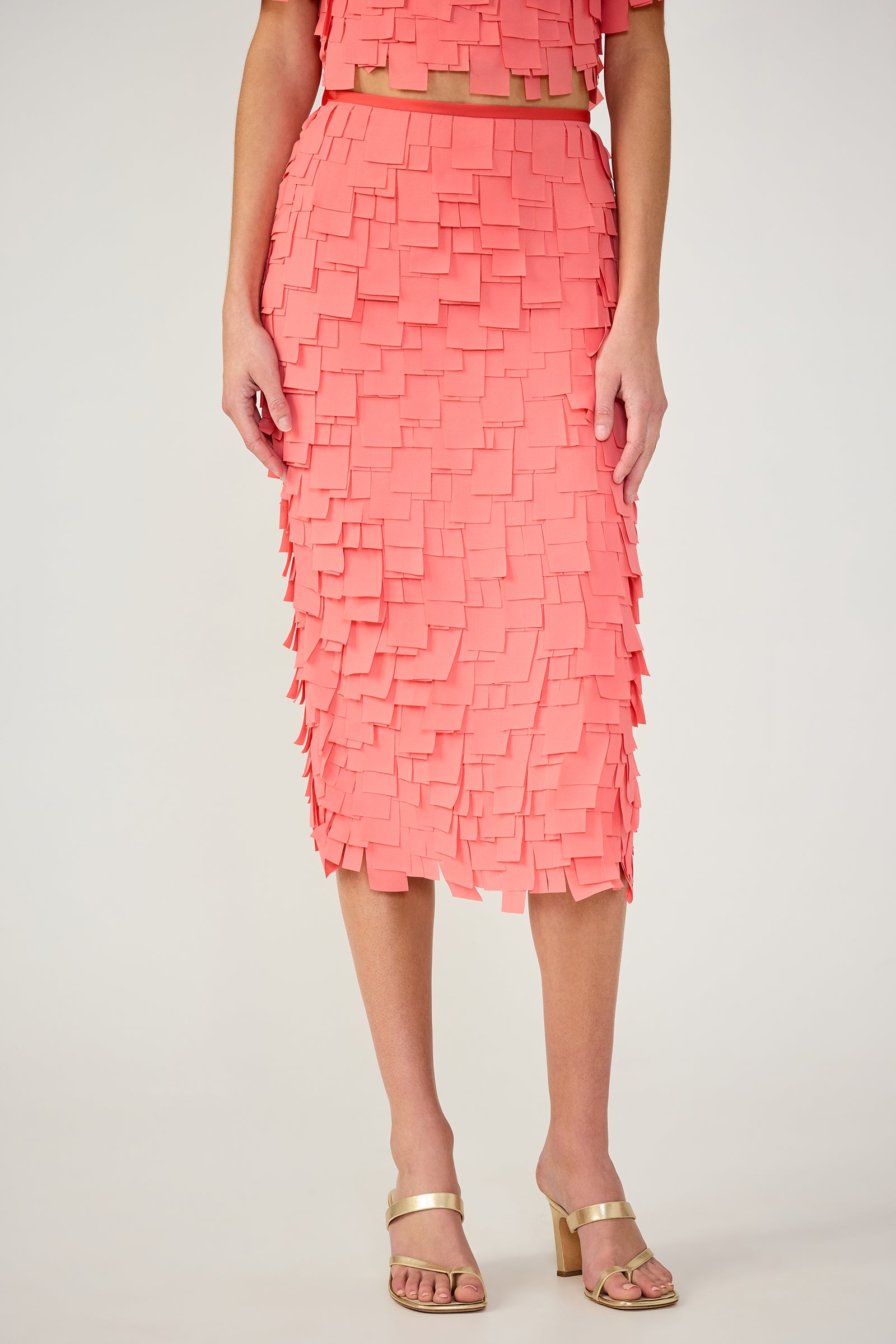 A woman wears the Dita Midi Pencil Skirt - Flutter Chiffon in Coral Crush, featuring layered square fabric pieces and a textured geometric pattern, falling below the knee. She pairs it with gold open-toe heels. Upper body partially visible.