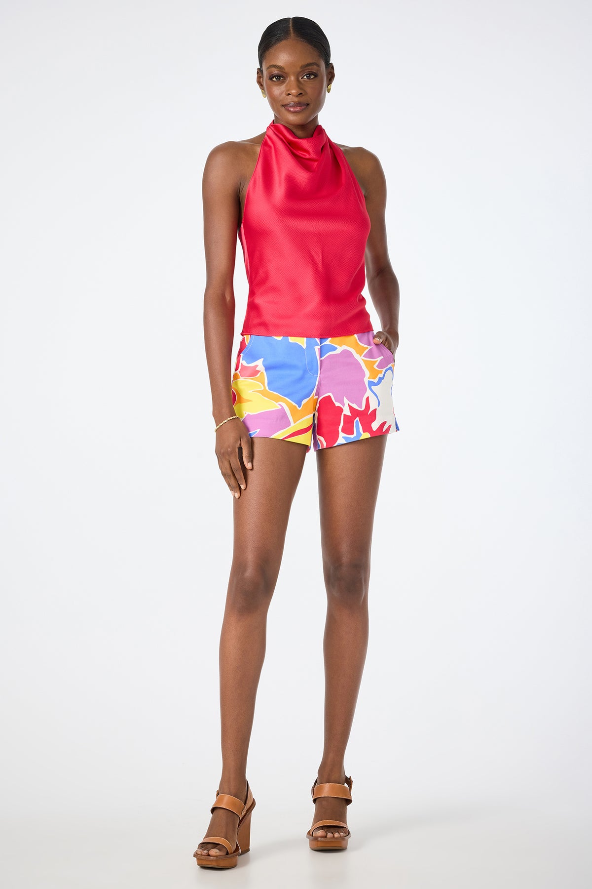 A woman smiles in the Corbin 2 Shorts - Soie Paradis, featuring colorful florals, paired with a sleeveless red halter top and tan heeled sandals against a plain white background.