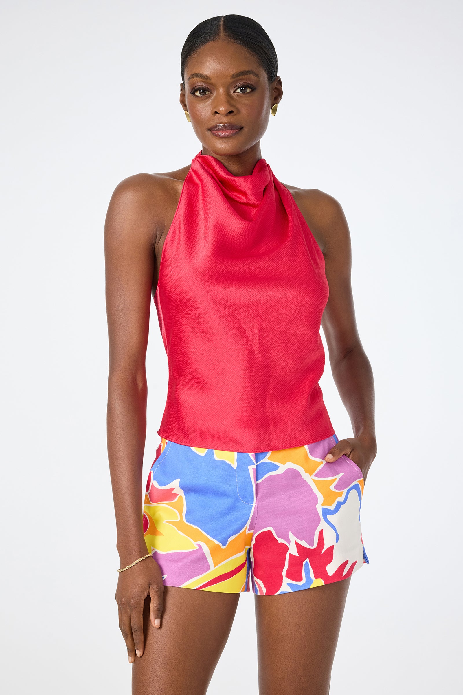 A woman wears the Lanika Halter Top in Cannes Coral, paired with colorful floral shorts, standing with one hand in her pocket against a plain white background.