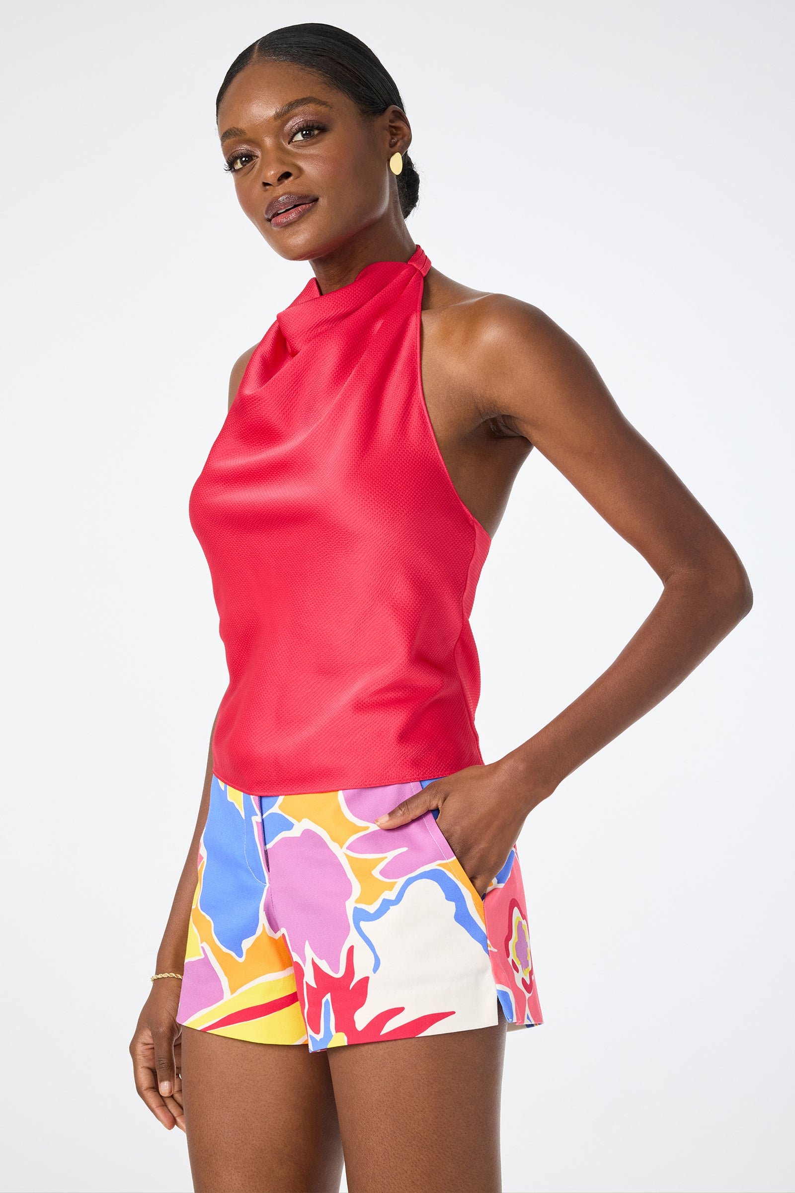 A woman poses confidently against a plain white background, wearing the Lanika Halter Top—a sleeveless, high-neck red halter—and colorful floral print shorts, with one hand in her pocket.