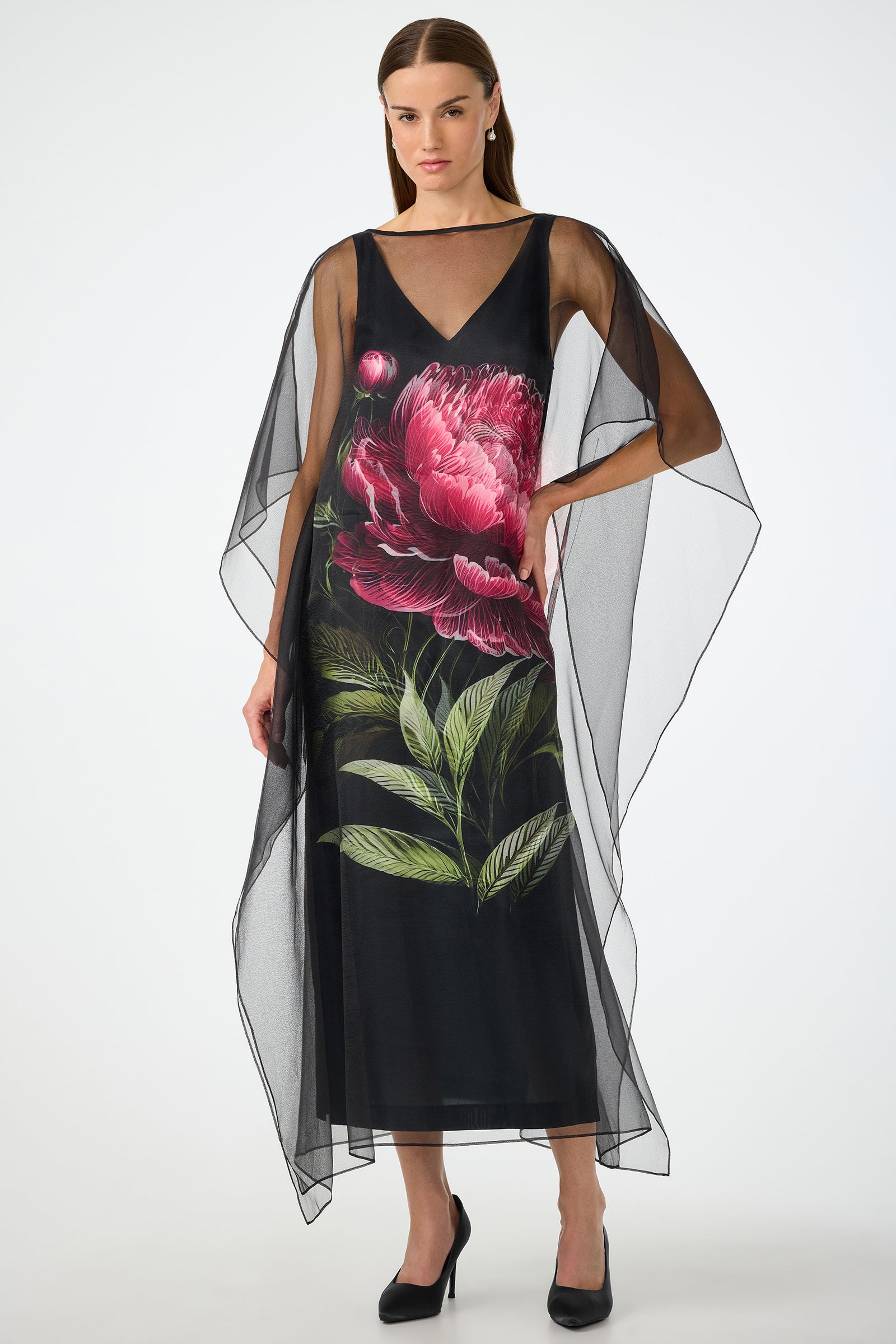 A woman wears the Salon 2 Organza Midi Dress in Fauna Floral, a sheer black cape dress with wide, flowing sleeves and bold pink peony and green leaf print on the front. She stands confidently in black heels with one hand on her hip.