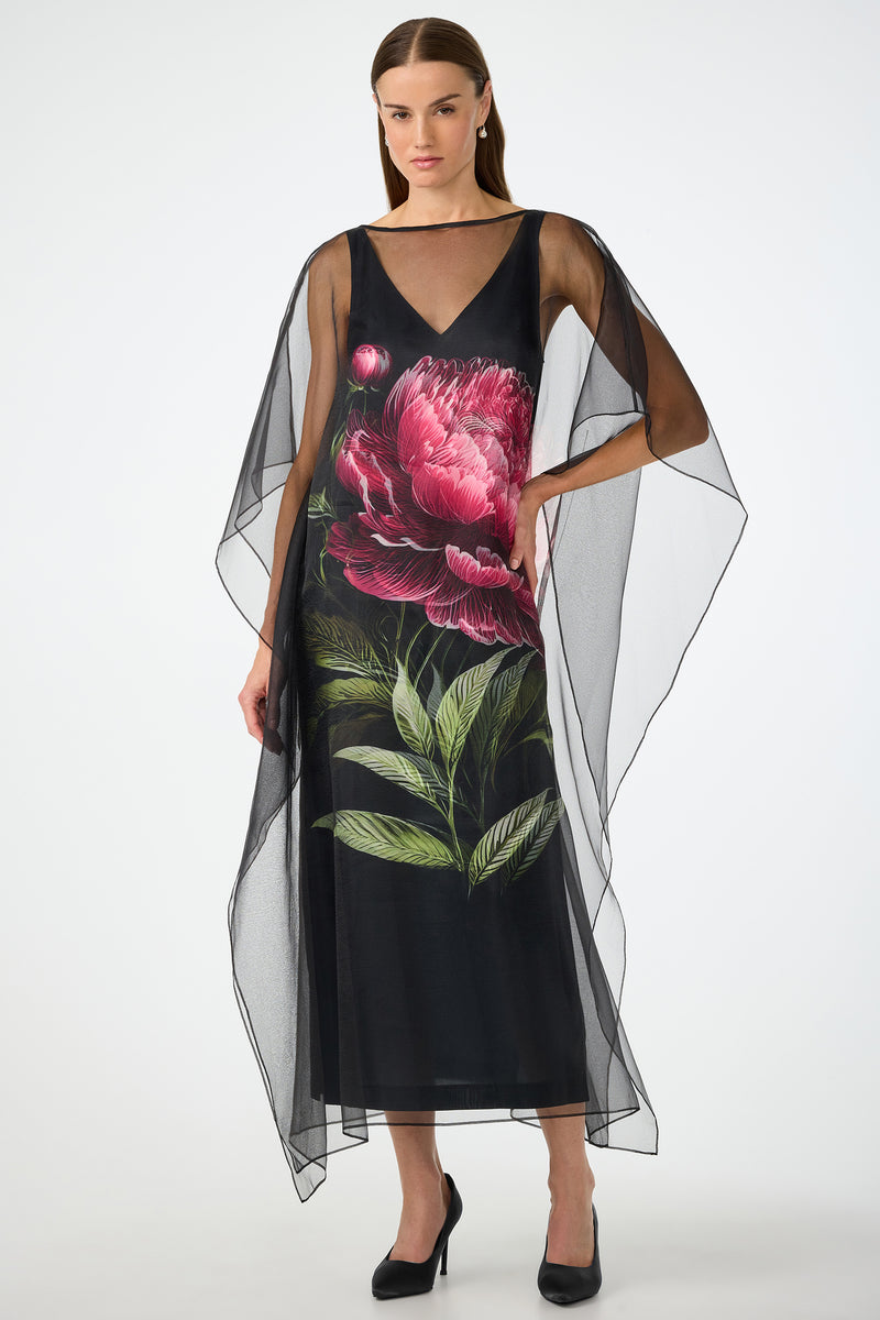 A woman wears the Salon 2 Organza Midi Dress in Fauna Floral, a sheer black cape dress with wide, flowing sleeves and bold pink peony and green leaf print on the front. She stands confidently in black heels with one hand on her hip.