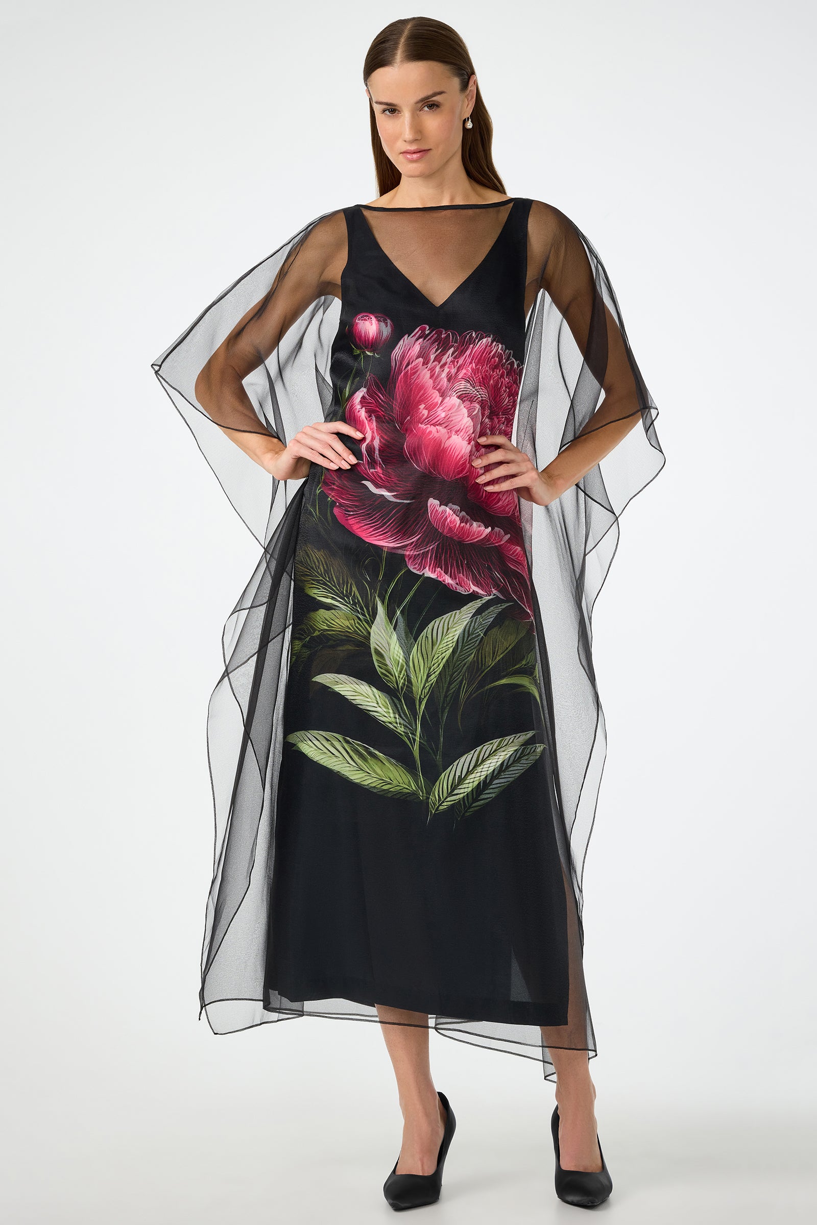 A woman poses with hands on hips in the Salon 2 Organza Midi Dress—a sheer black midi featuring wide, flowing sleeves and a bold pink flower with green leaves printed on the front. She completes the look with black heels.