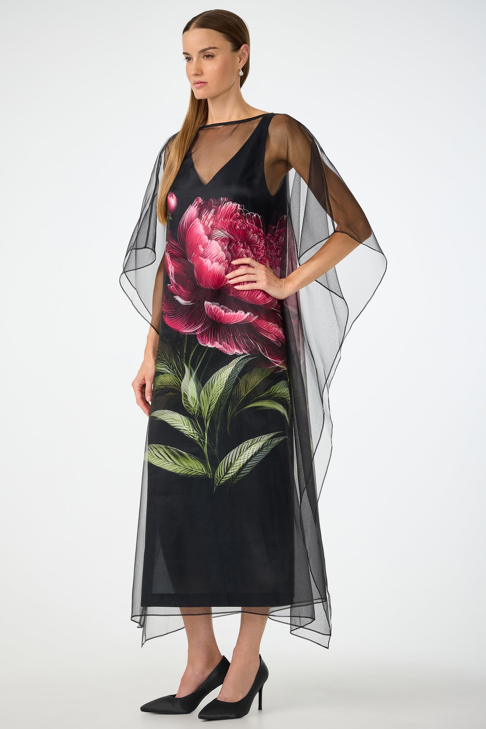 A woman stands in profile, hands on hips, wearing the Salon 2 Organza Midi Dress— a long black dress with sheer overlay and large pink flower with green leaves—paired with black heels against a white background.