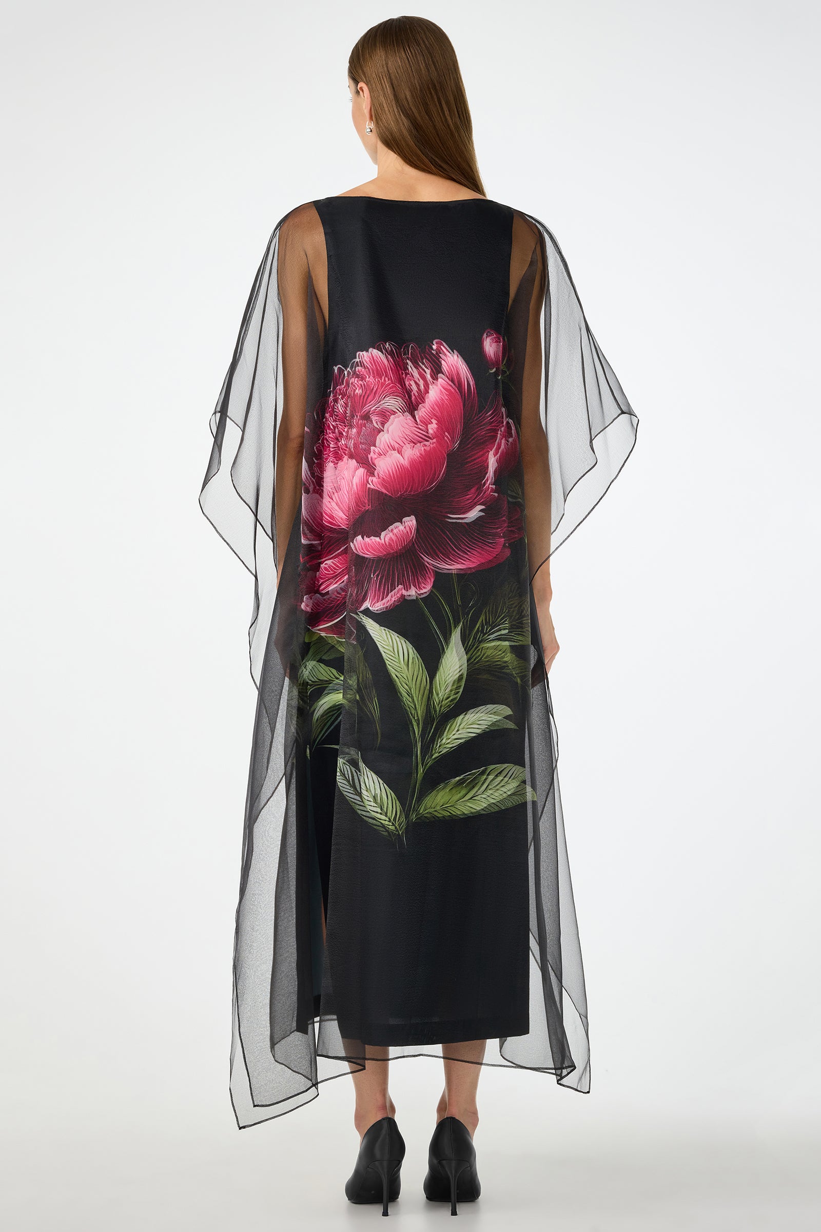 A woman faces away, wearing the Salon 2 Organza Midi Dress—a long black sheer dress with flowing sleeves and a large vibrant pink flower with green leaves on the back. She has straight brown hair and black shoes.