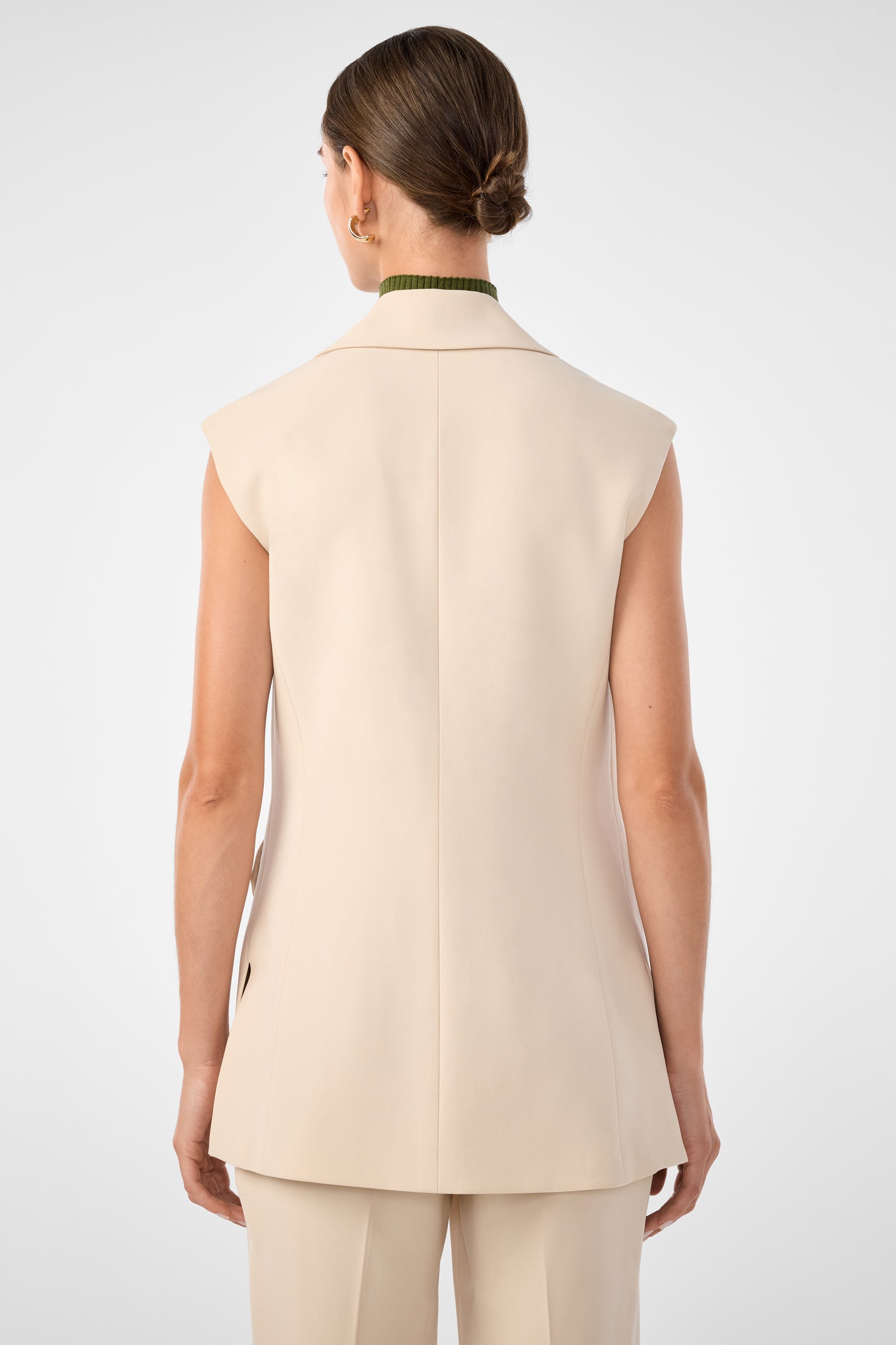 A woman faces away, wearing the Boabab Sleeveless Jacket in beige with matching trousers. Her hair is styled in a low bun, showing a green collar above the jacket. The backdrop is plain and light-colored.