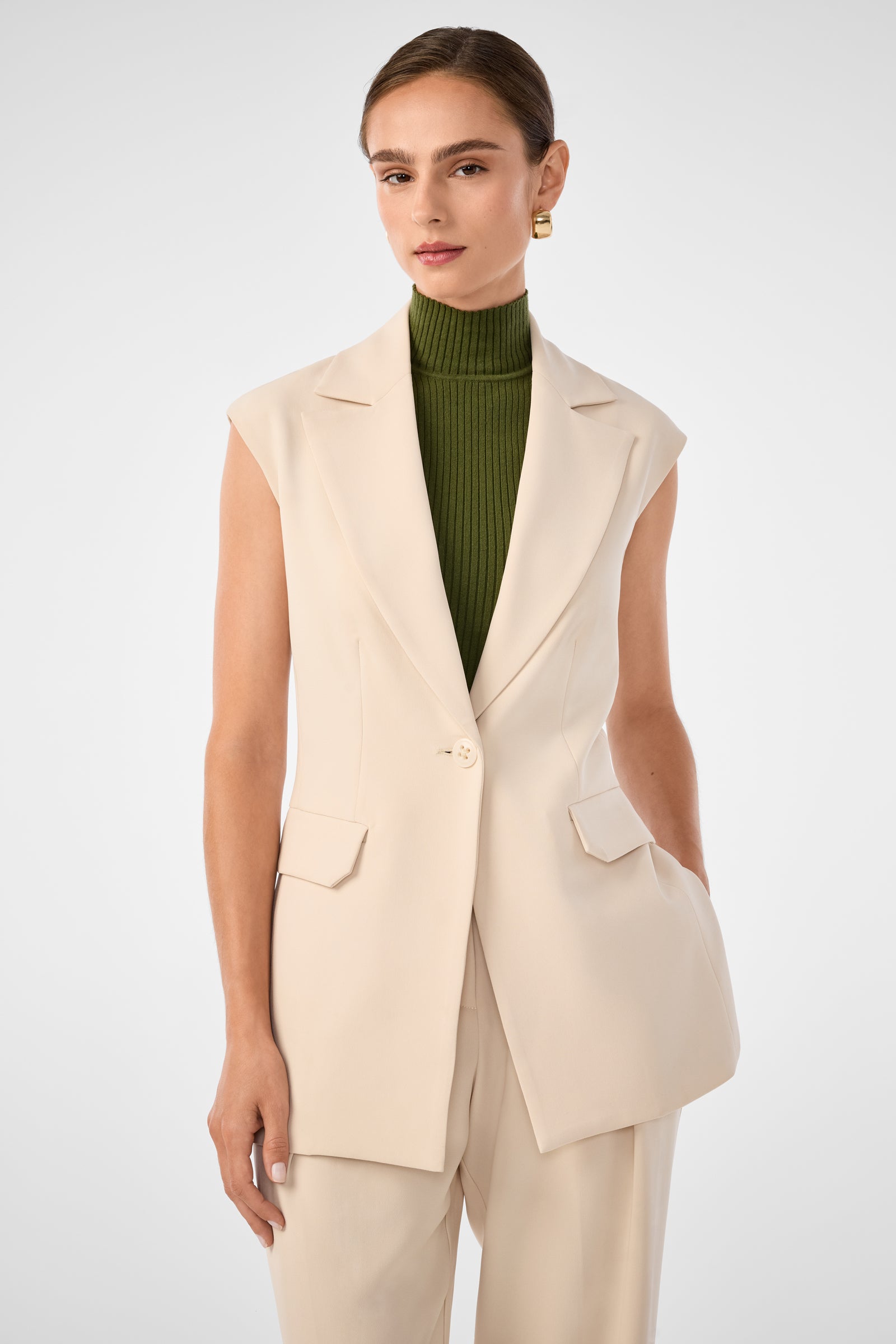 A woman wears the Boabab Sleeveless Jacket in Poplar over a ribbed olive green turtleneck and matching beige pants, standing against a plain light gray background.