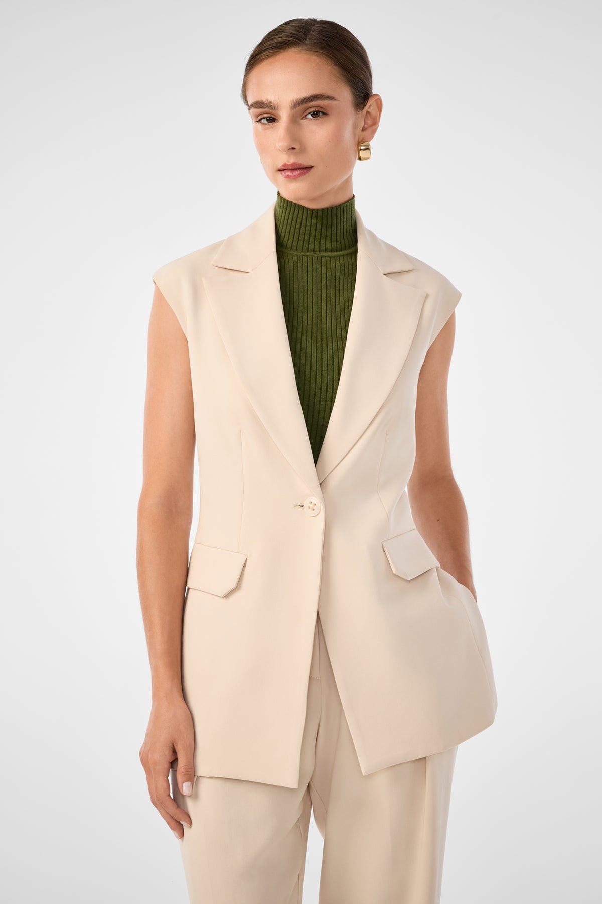 A woman wears the Boabab Sleeveless Jacket in Poplar over a ribbed olive green turtleneck and matching beige pants, standing against a plain light gray background.